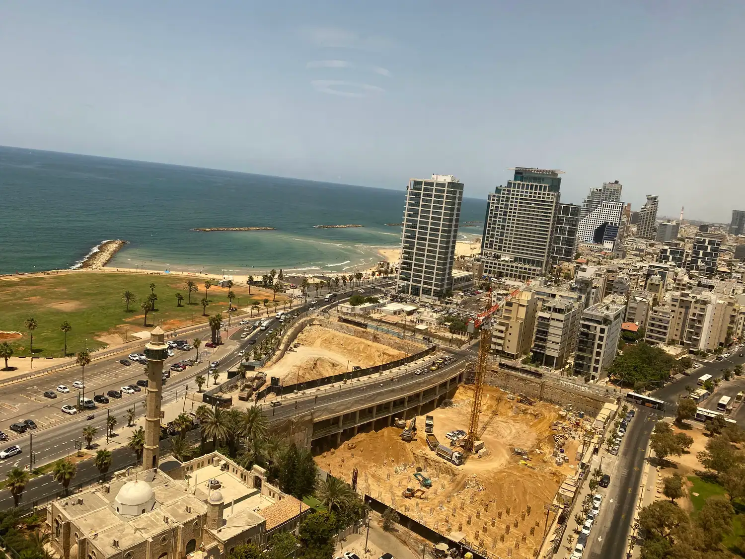The coast seen from high above: the sea and a small harbour, a stone building with a minaret and a dome, the beach and its lawns, a palm lined road, and tower blocks beyond a building site.