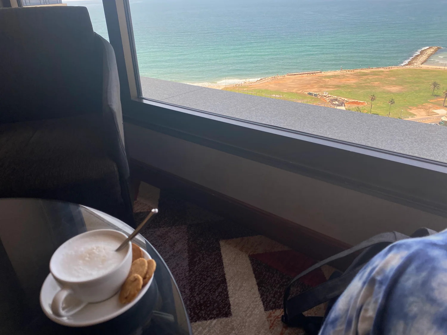A cup of coffee and two biscuits on a glass table beside a window, with the sea and a strip of green shoreline beyond the glass.