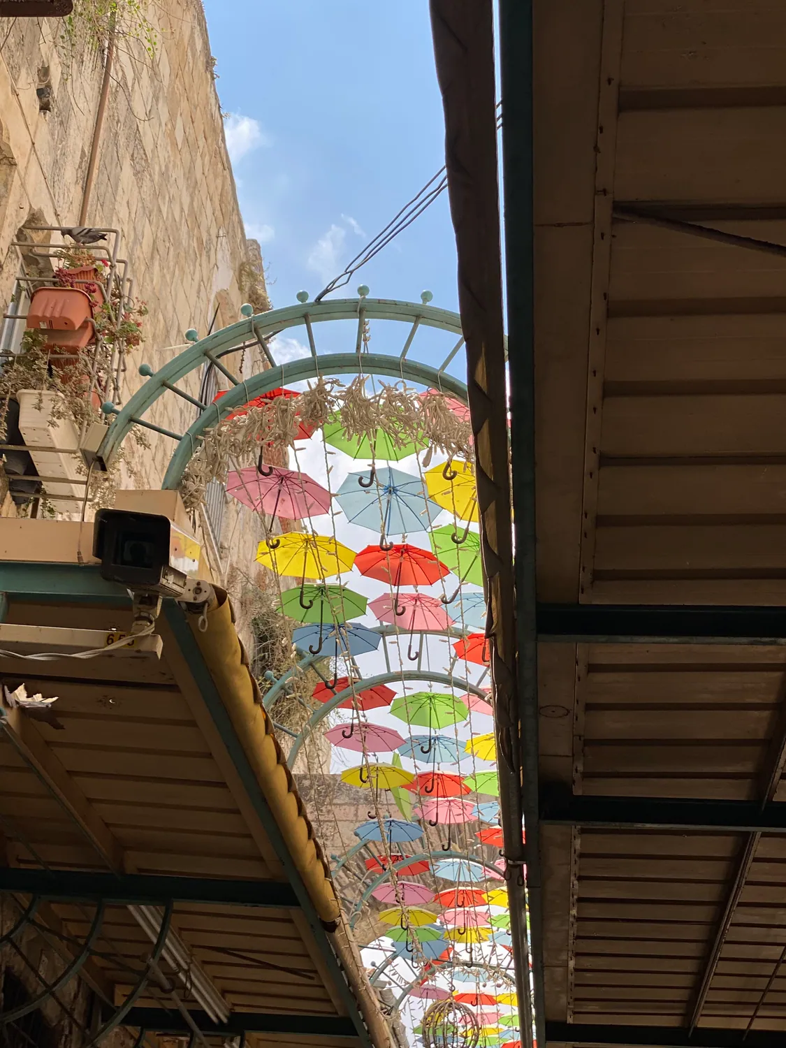Looking up at rows of bright umbrellas strung overhead between the stone walls of a covered lane, with blue sky above them.