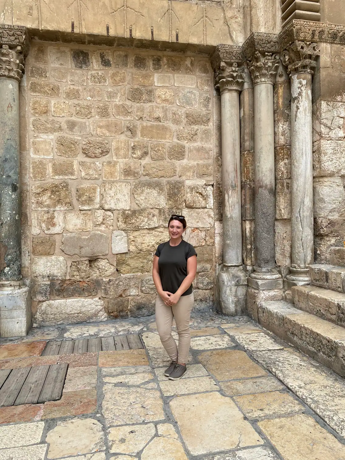 A woman in a dark top and light trousers standing on flagstones in front of an old stone wall and carved columns.