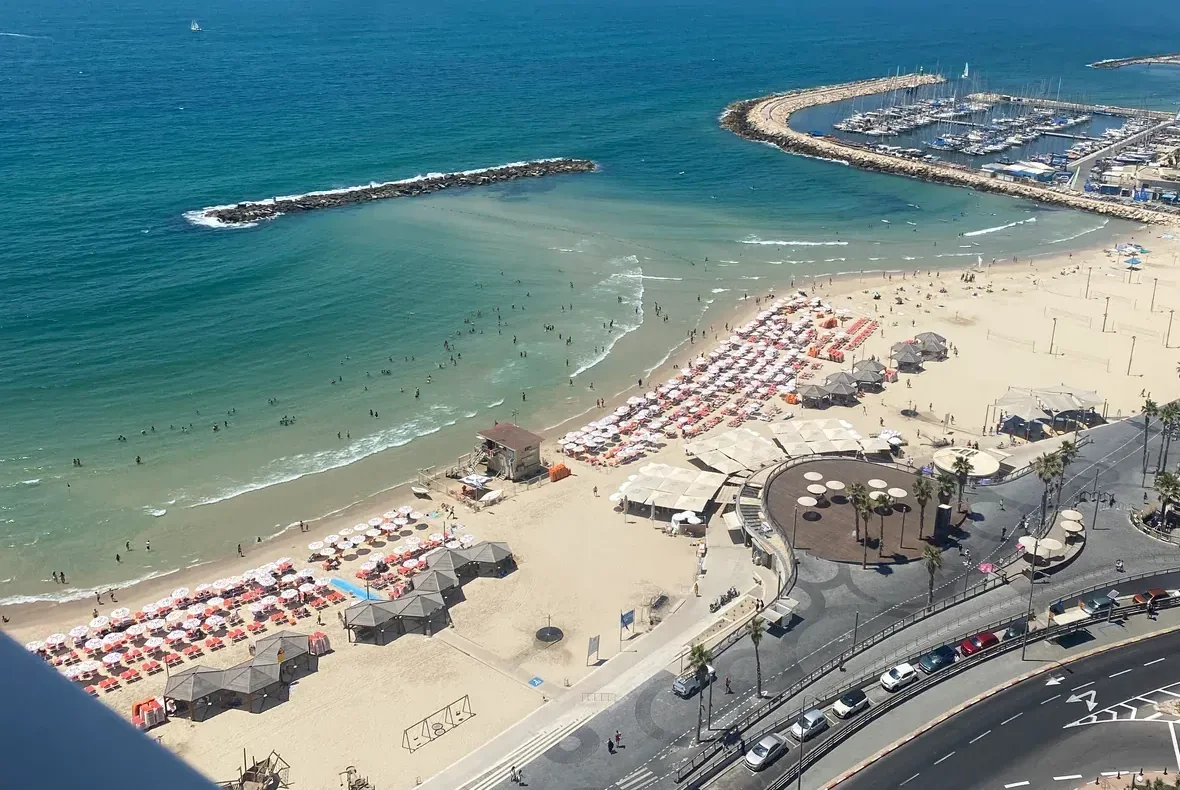 A beach from above on a bright afternoon: rows of sun umbrellas and low beach shelters on the sand, a marina of small boats inside a curved breakwater, the sea beyond, and a seafront promenade along the bottom of the frame.