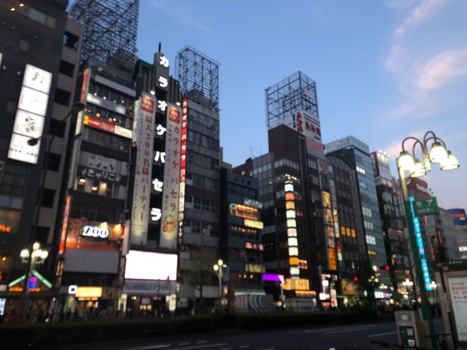 A cluster of lit buildings at dusk, their faces lined with signs and a street lamp standing at the right, against a deep blue sky.