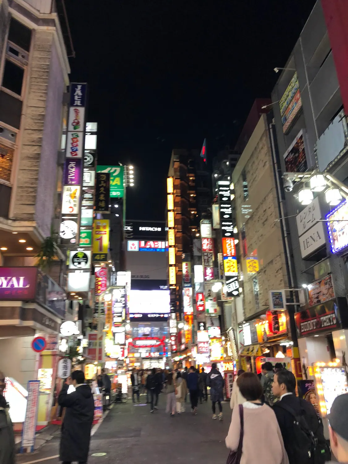 A night street of signs with people walking between the shopfronts, their lights on along both sides.