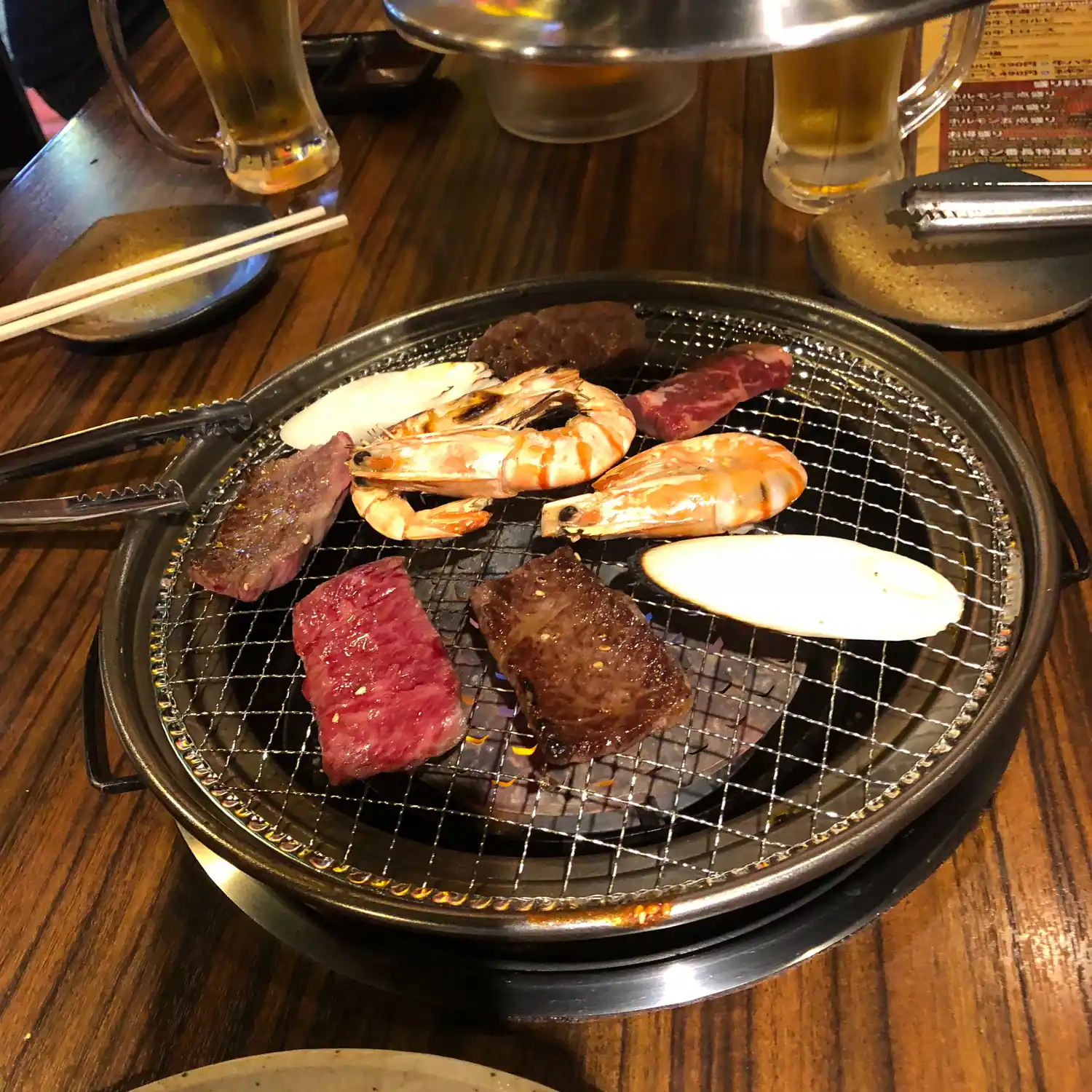 Meat and seafood grilling on a table grill, with chopsticks and a glass beside it.