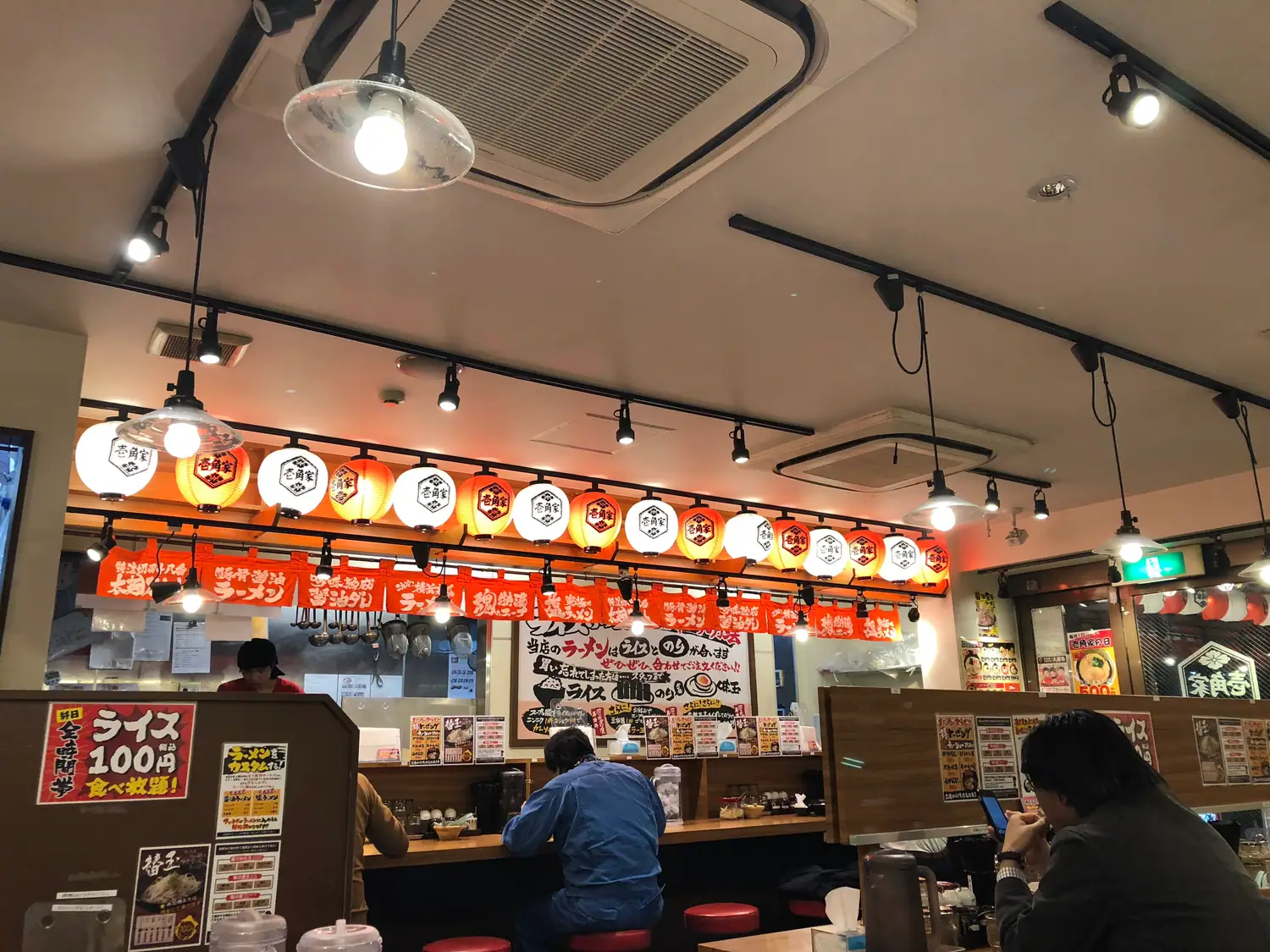 A small counter bar with paper lanterns hung along the counter, diners seated on stools and printed menus on the wall behind them.