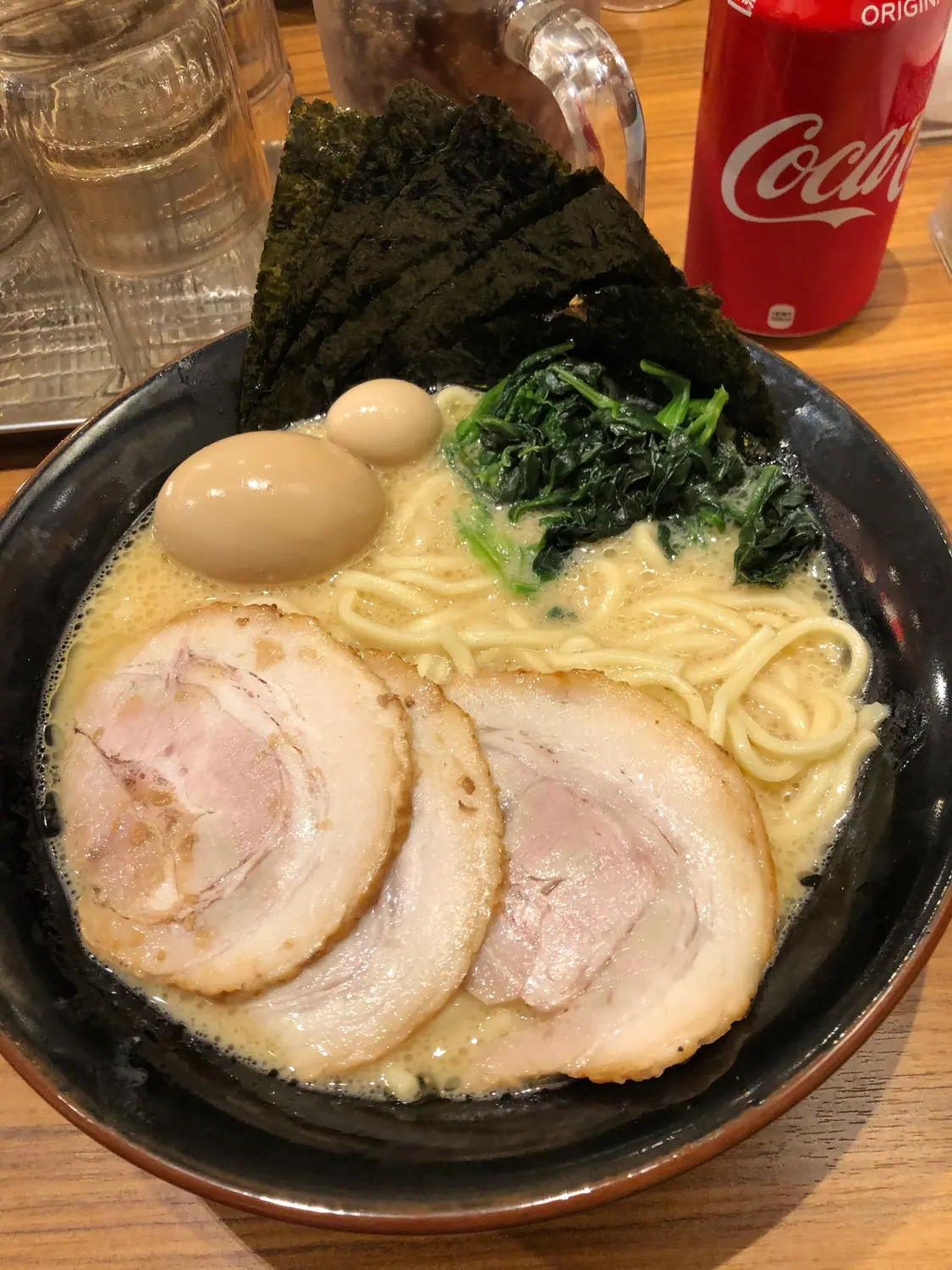 A bowl of noodles with sliced pork, egg and greens, a can standing beside it on the counter.