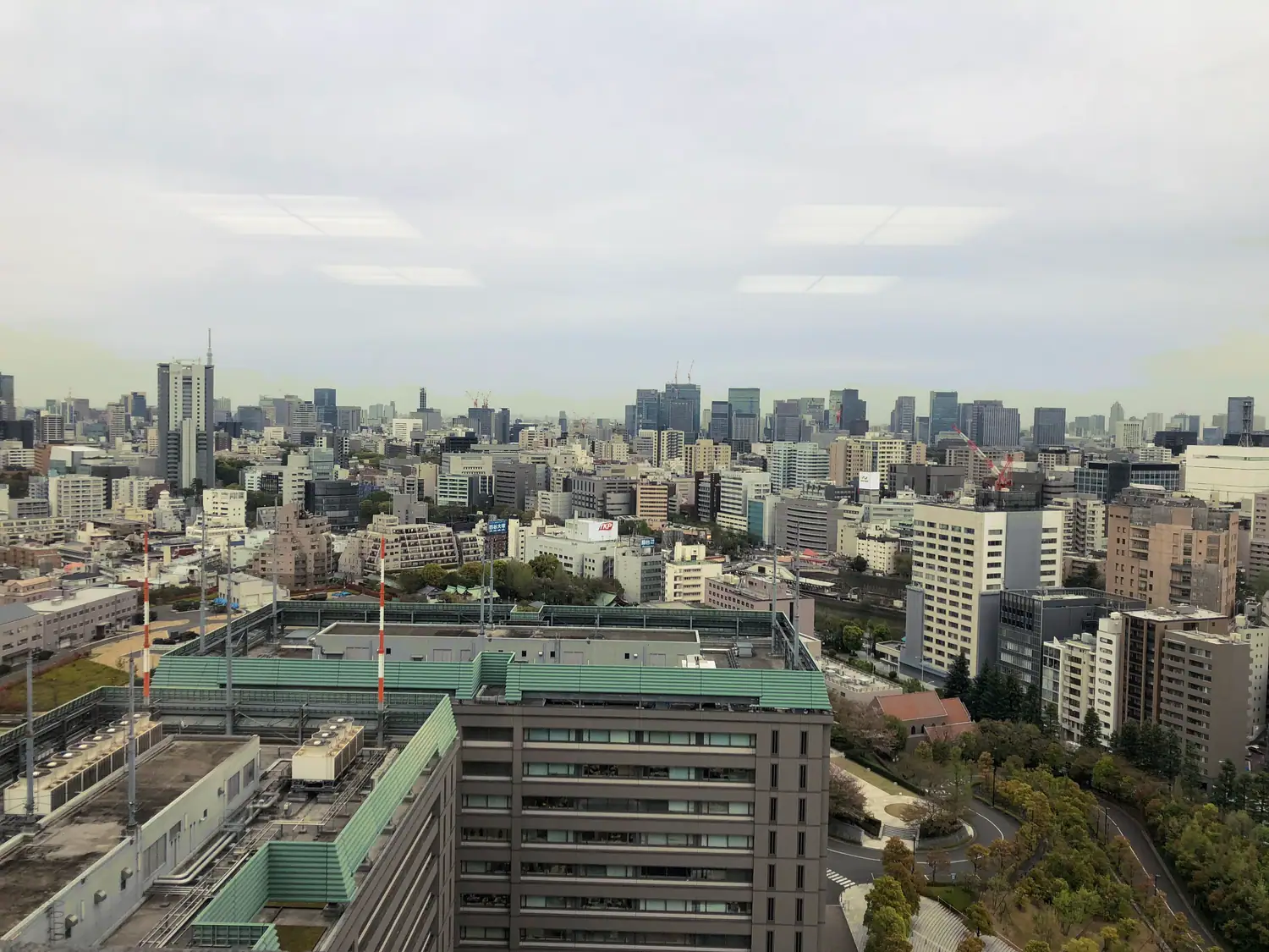 A city seen from above with a green park and low rooftops, taller towers along the horizon under a hazy sky.