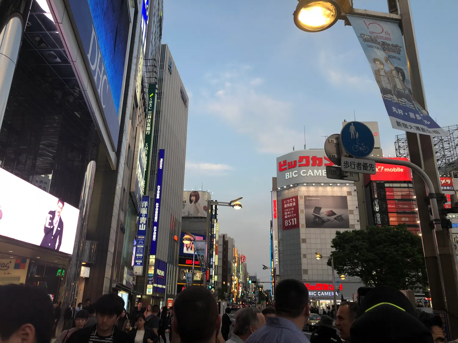 A street of shops at dusk with screens lit above the crowd, people walking between the shopfronts.