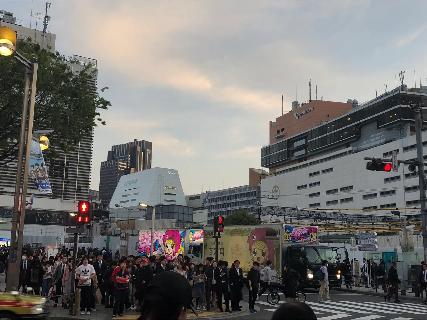 A crossing filled with people at dusk, a pale building with a lit screen across the street and lamps strung overhead.