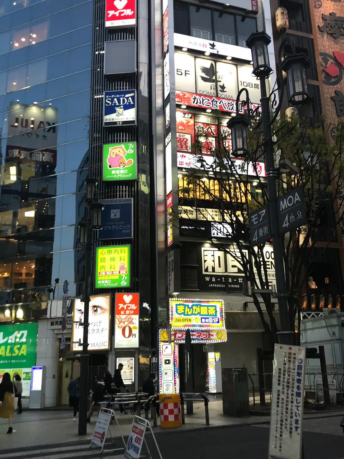 A street of lit signs with a few people at the edge of the frame, shopfronts down both sides.