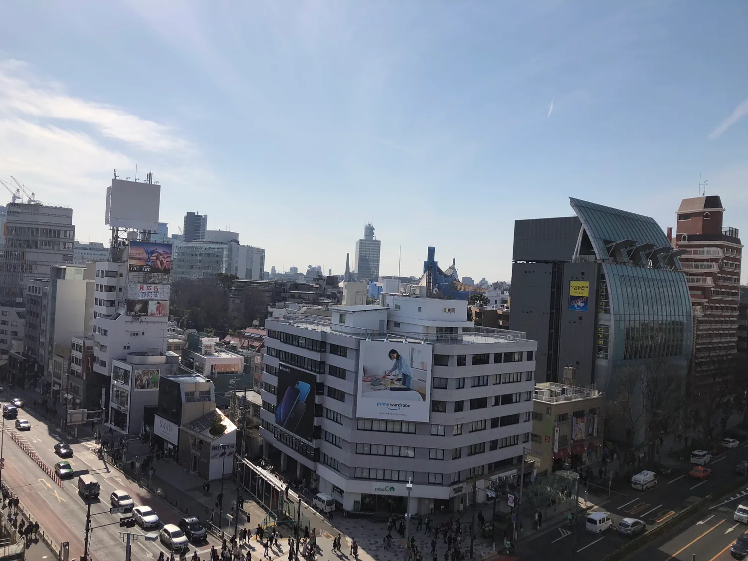 A city seen from above between tall buildings: a wide street of traffic and crossings below, rooftops and towers packed to the horizon under a pale blue sky.