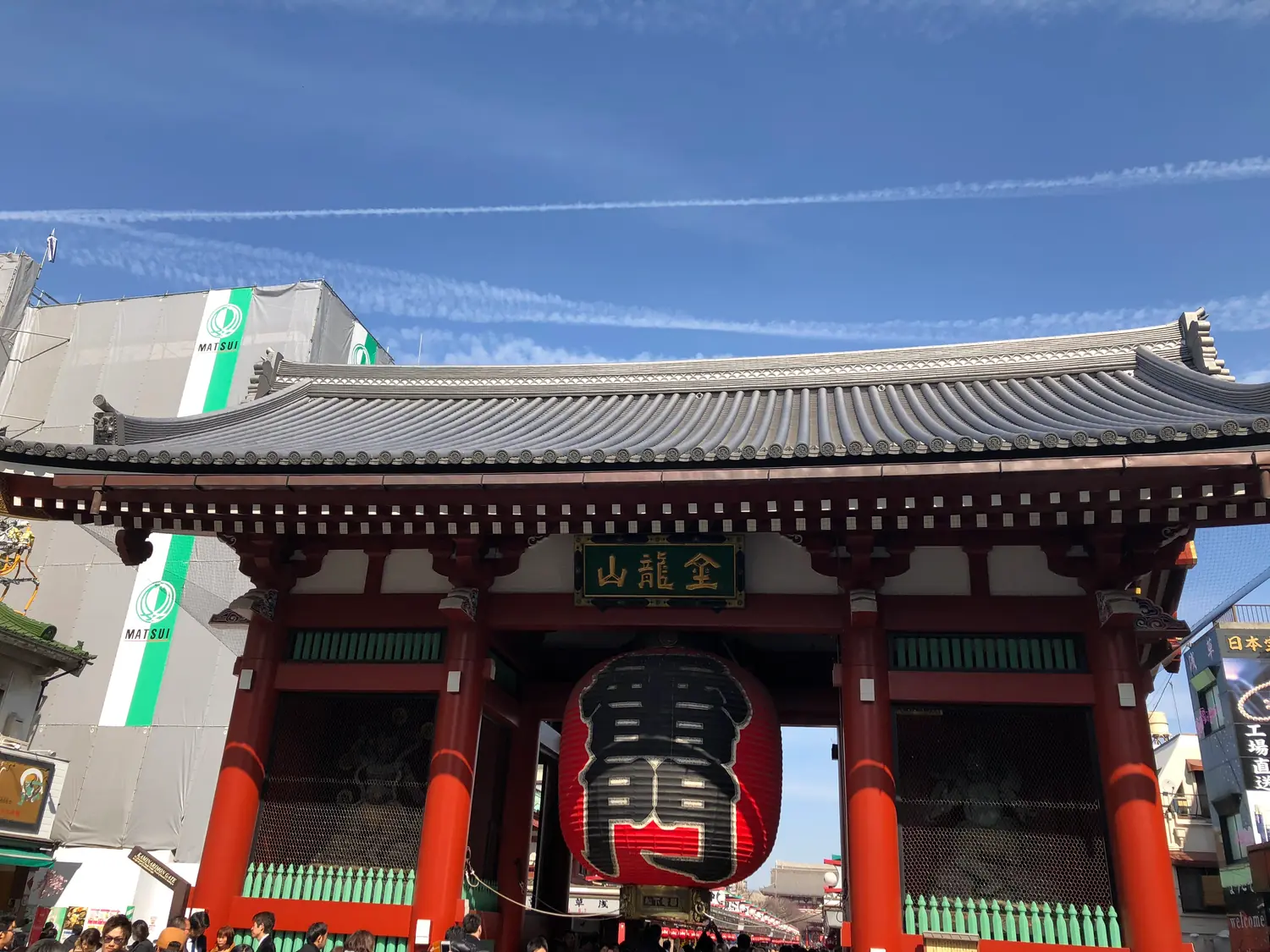 A tall temple gate with red pillars and a wide green tiled roof standing against a clear blue sky, a great red paper lantern hanging at its centre and low buildings and trees at either side.