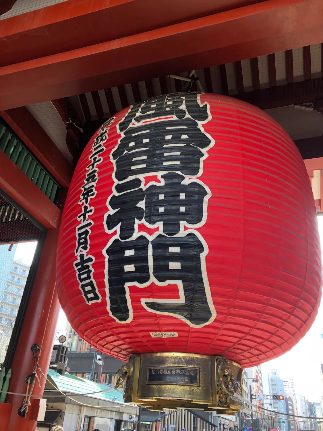 A great red paper lantern filling the frame, its paper lettered in dark characters, hanging under the heavy red beams of a gate.
