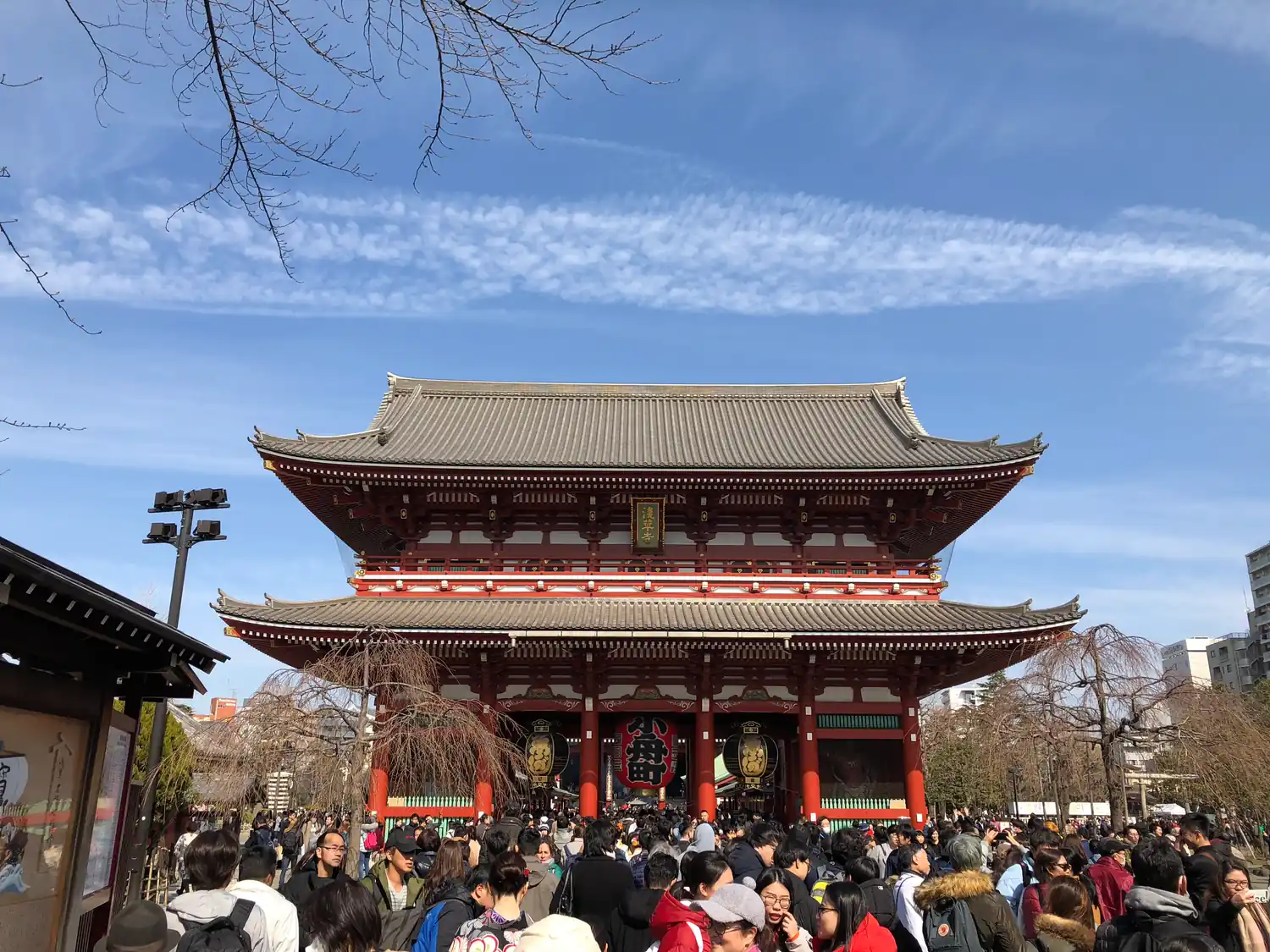 A wide temple hall with a green tiled roof and red pillars against a blue sky, a crowd of visitors gathered in front of it.