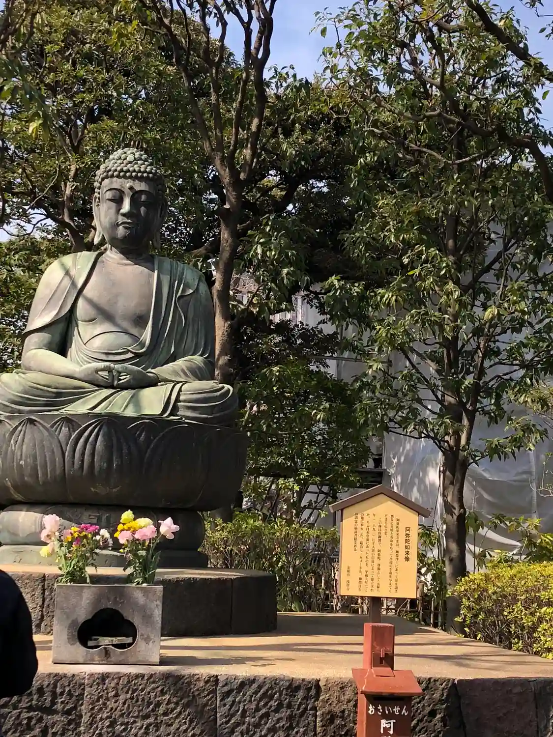 A seated bronze figure in a garden courtyard, a low stone lantern and clipped shrubs around it and bare trees behind.