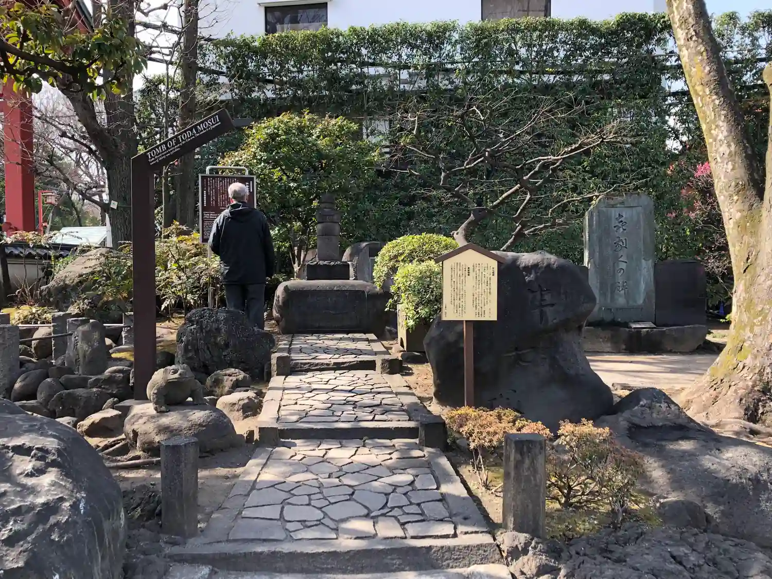 A stone path through a garden between trimmed shrubs and stone markers, a visitor in a dark coat walking away up the path ahead.