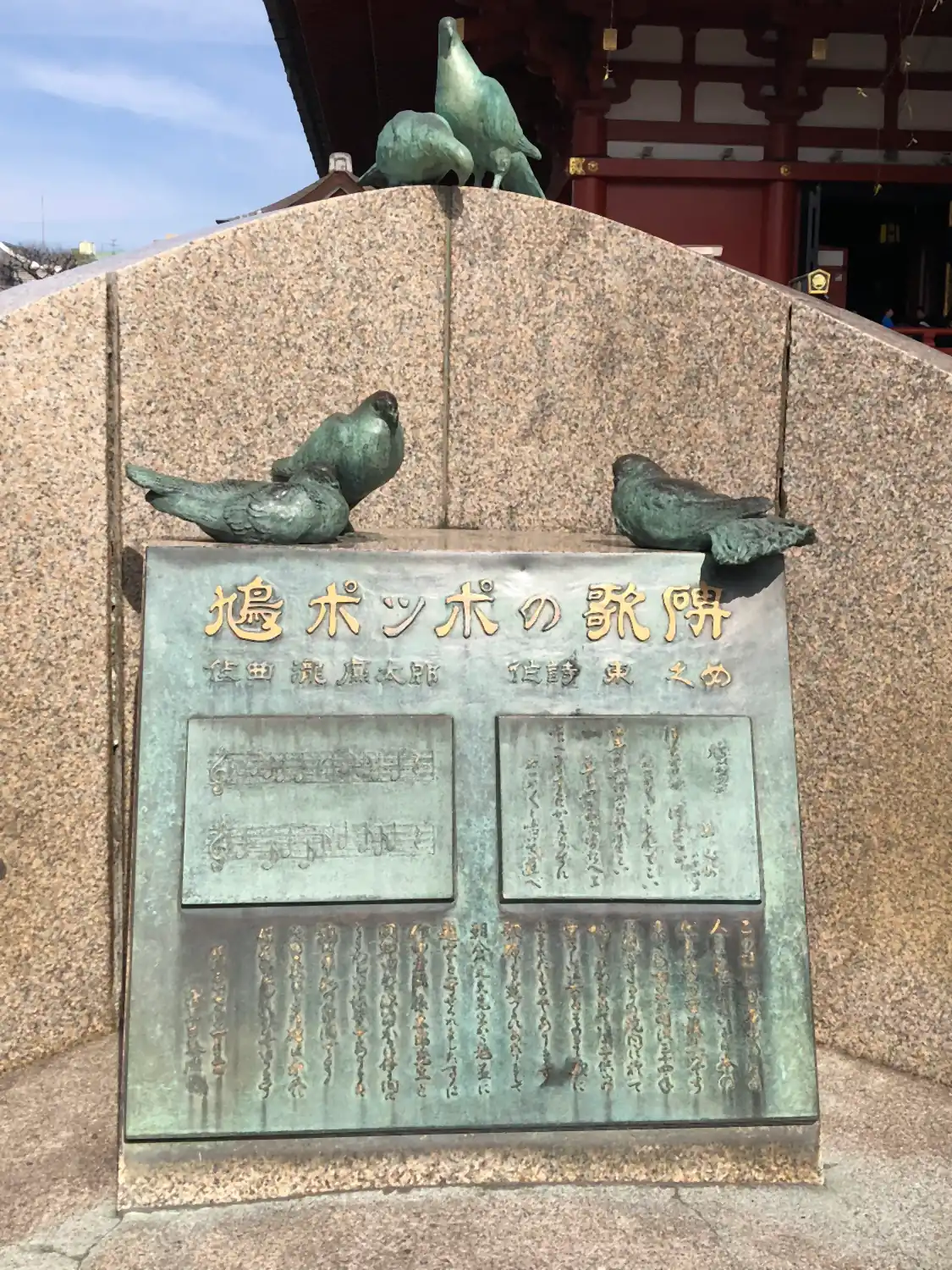 A granite monument with a bronze plaque in relief, four bronze pigeons perched on its ledges and a temple roof and blue sky above it.