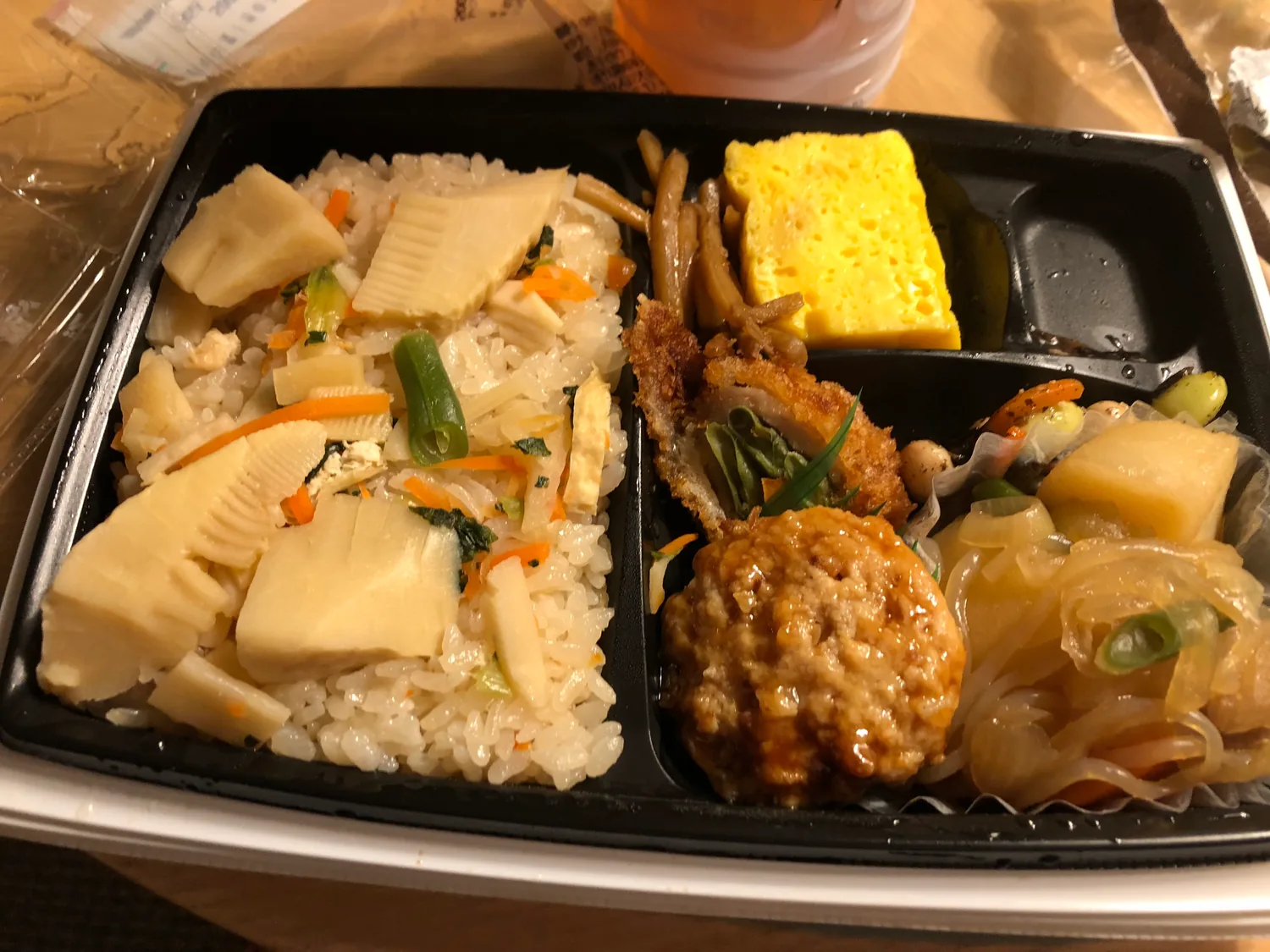 A boxed meal of rice with bamboo shoots, a rolled omelette, a croquette, a meatball and vegetables in a divided tray on a table.