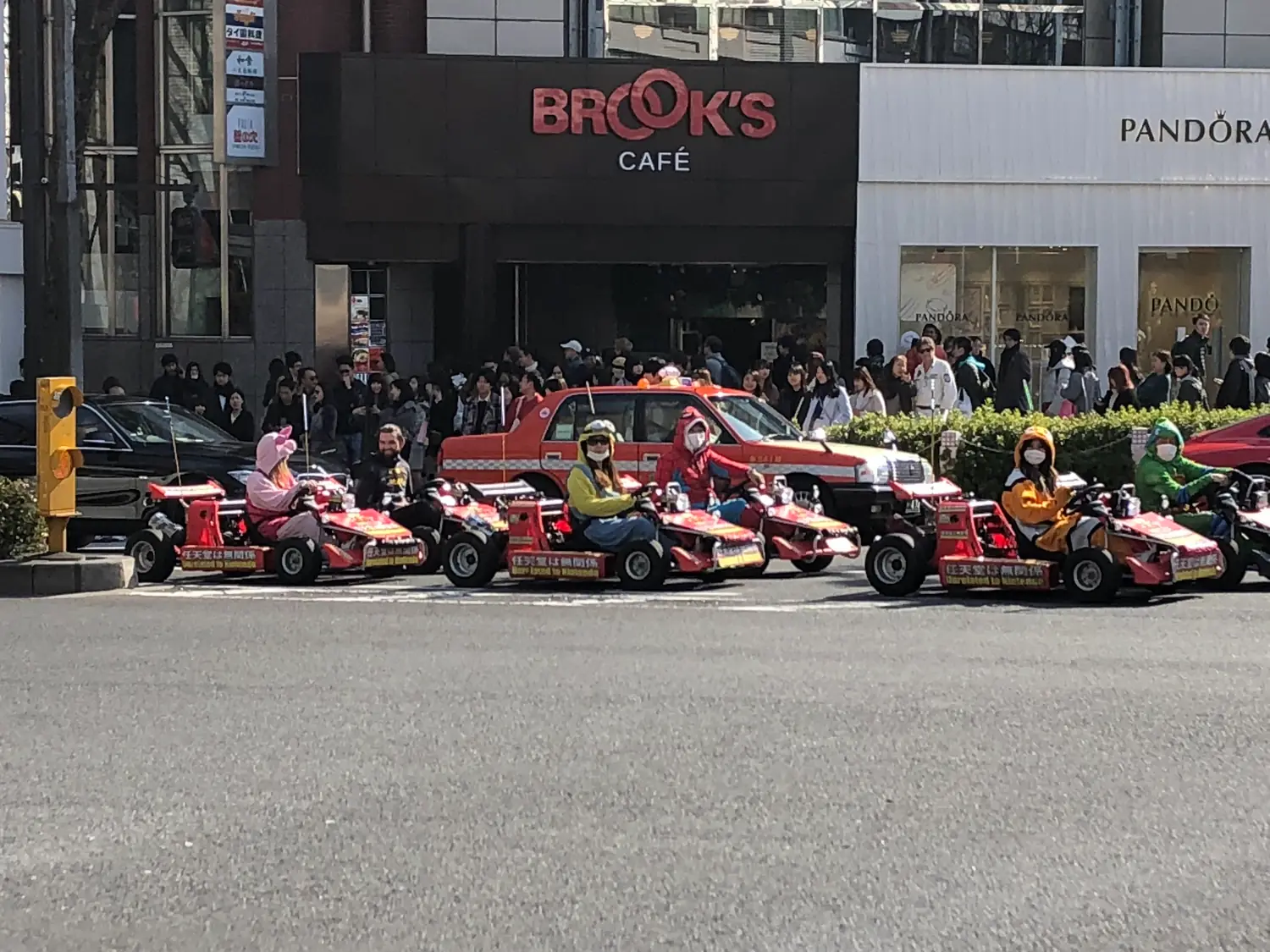 Small open karts with costumed drivers queued along a city street, shopfronts and a crossing beyond them.