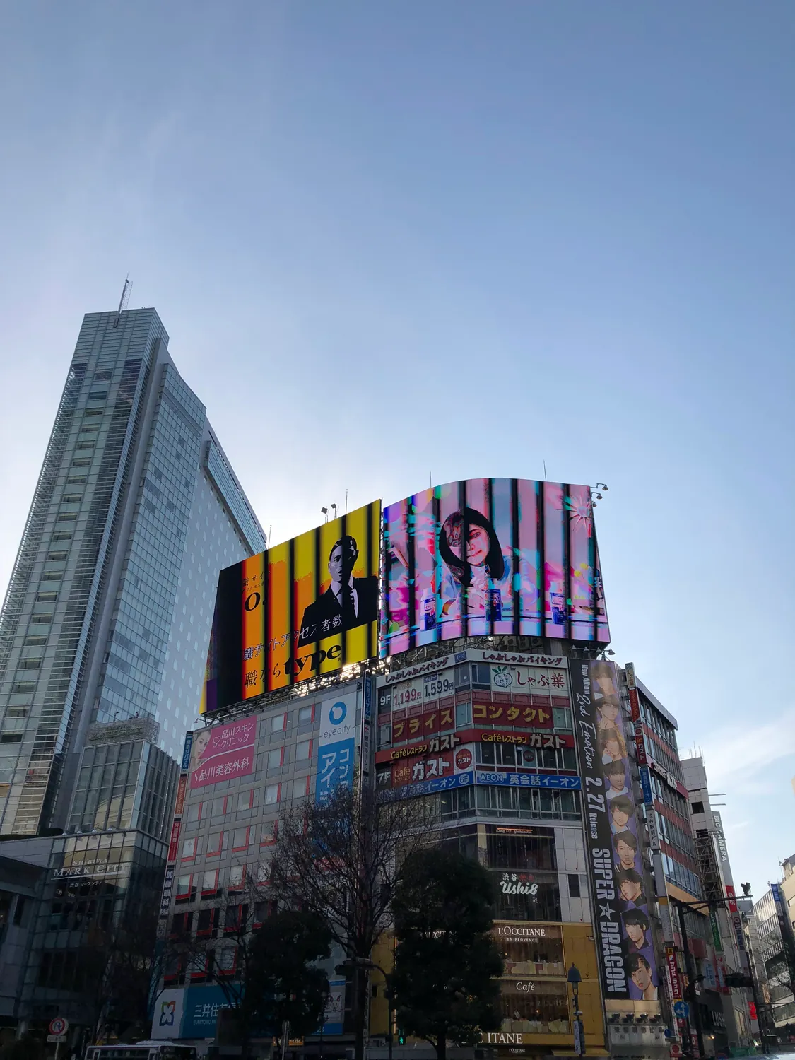 A tall building with a screen and a billboard on its face rising above lower blocks against a blue sky.