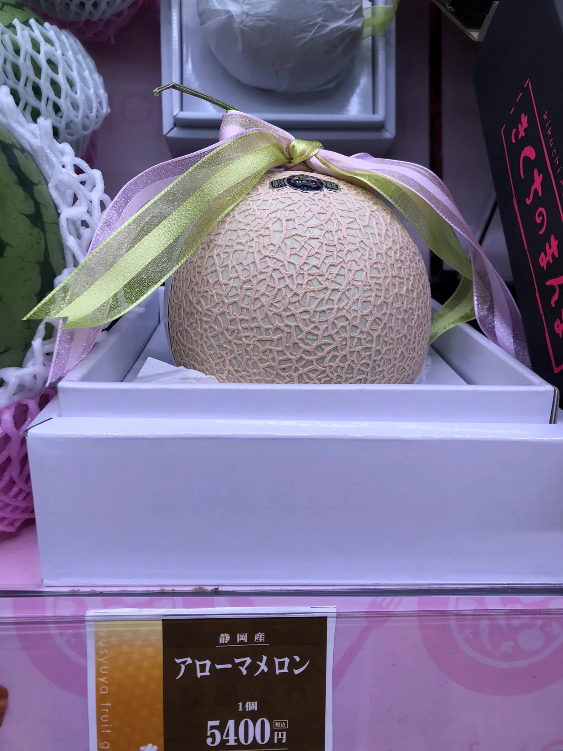A round melon in a presentation box with a ribbon, a price card standing in front of it.