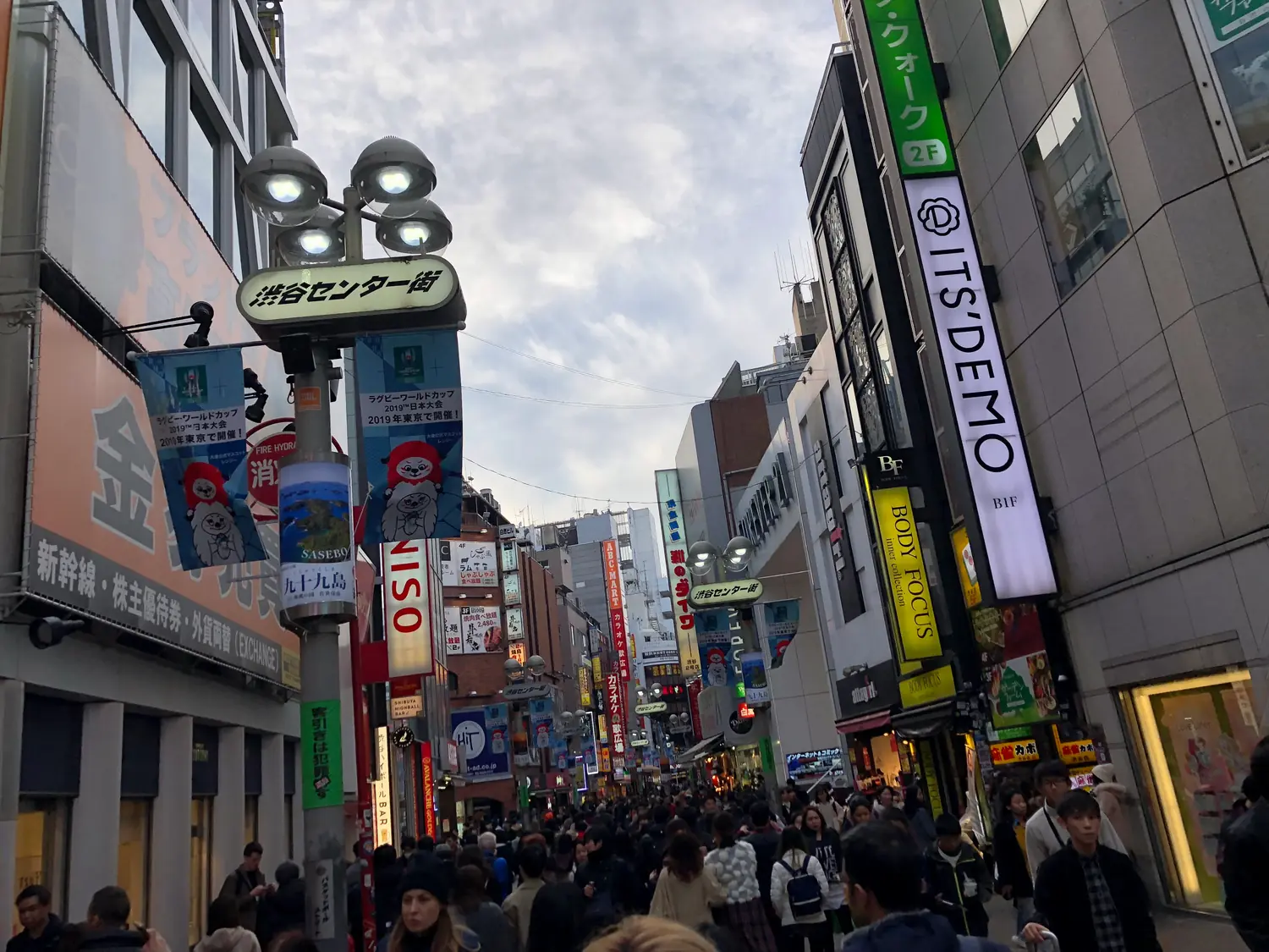 A narrow lane packed with people, tall vertical signs hanging on both sides and a grey sky above the roofs.