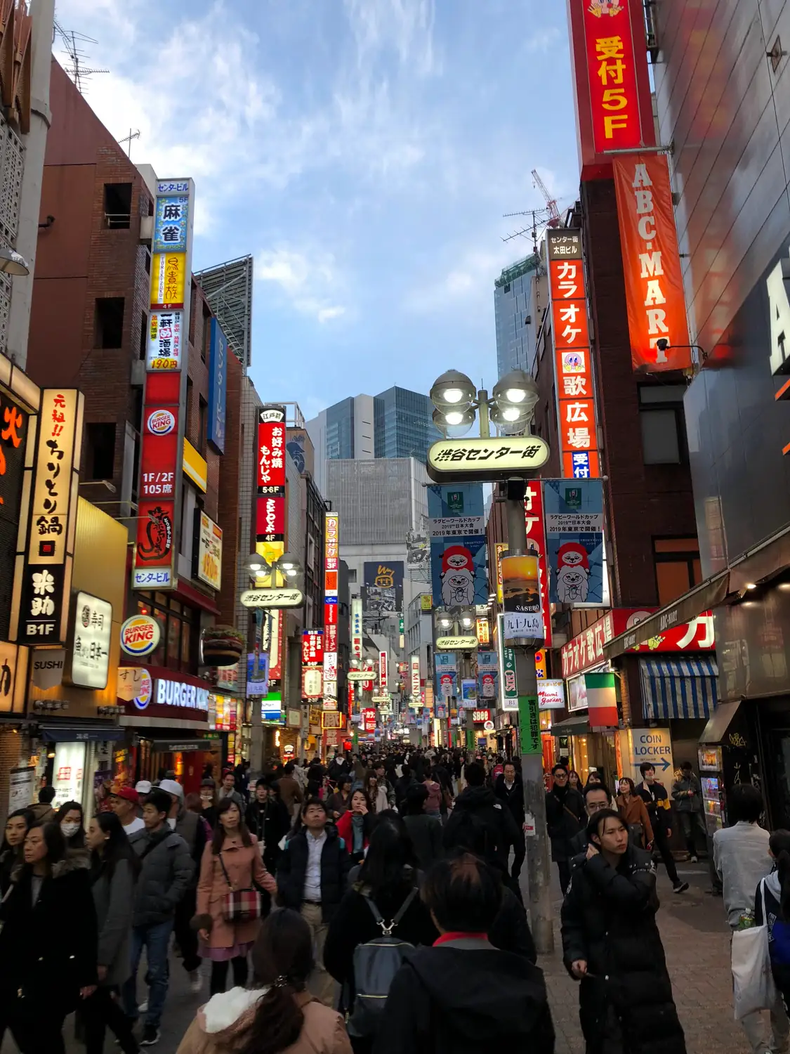 A crowded street at dusk, the signs lit along the shopfronts and the lane filled with people.
