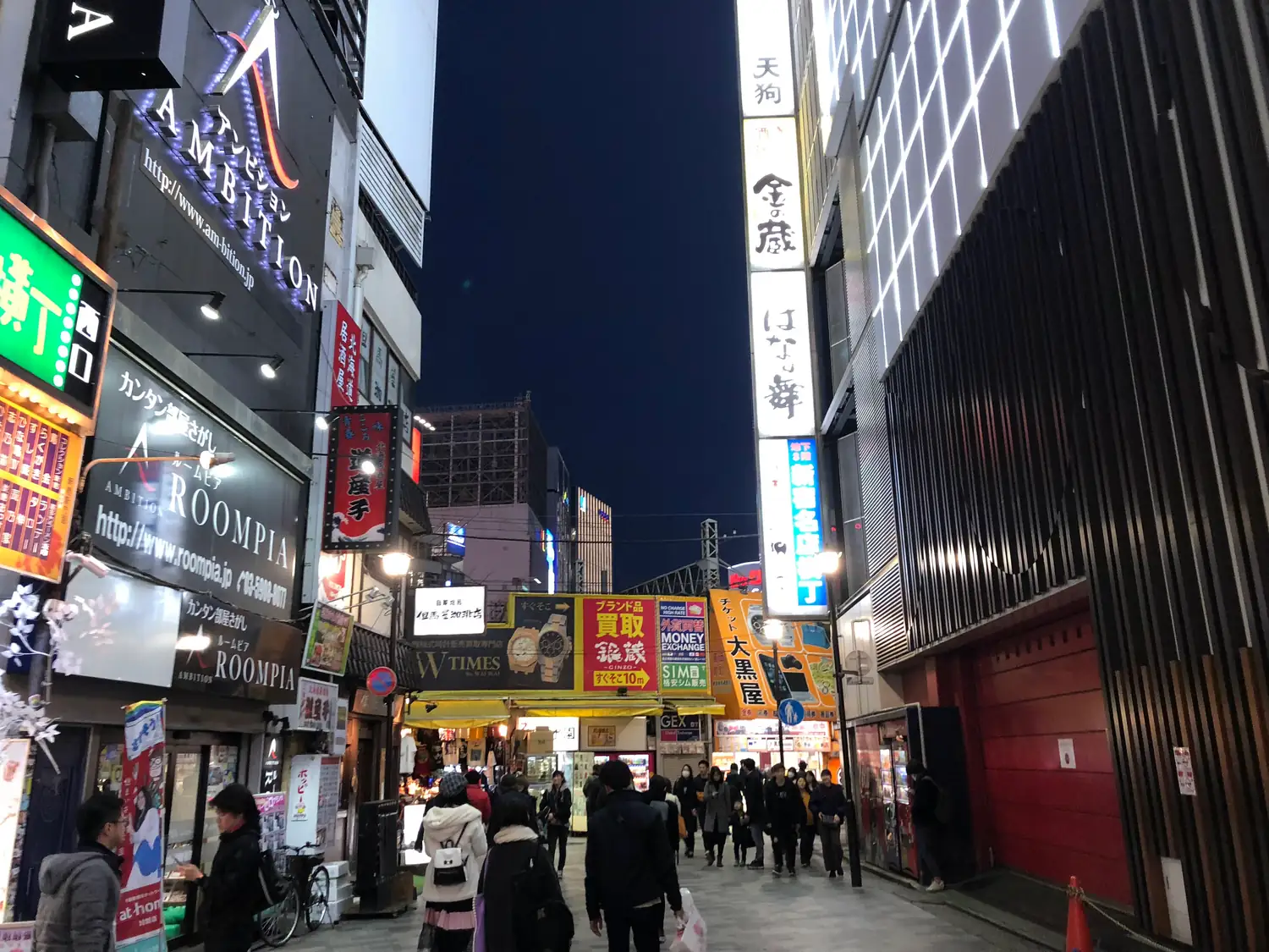 A lit street at night with a few people walking away between the shopfronts.