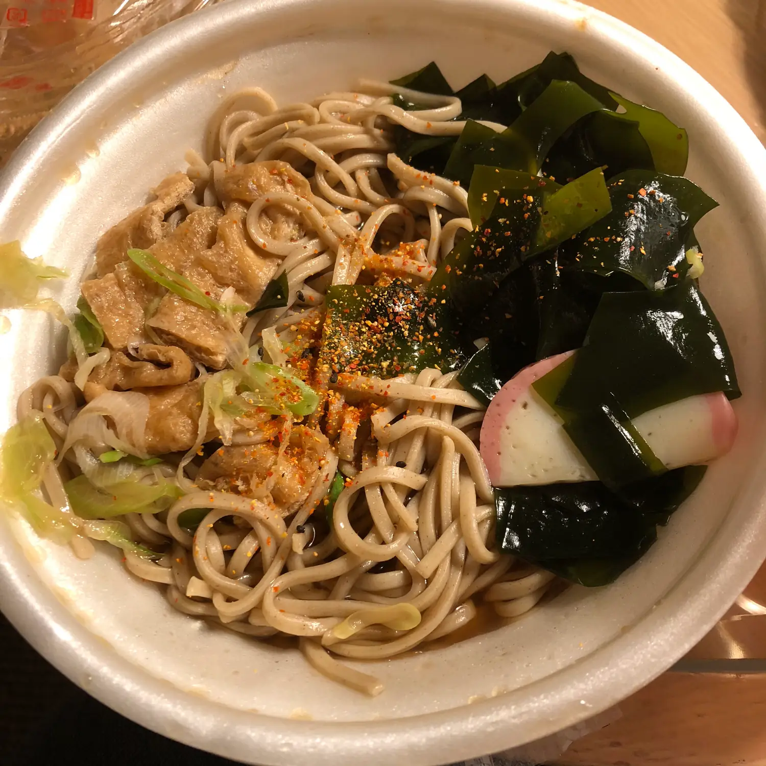 A bowl of noodles with seaweed, sliced pork and a slice of pink and white fish cake, dusted with chilli powder.