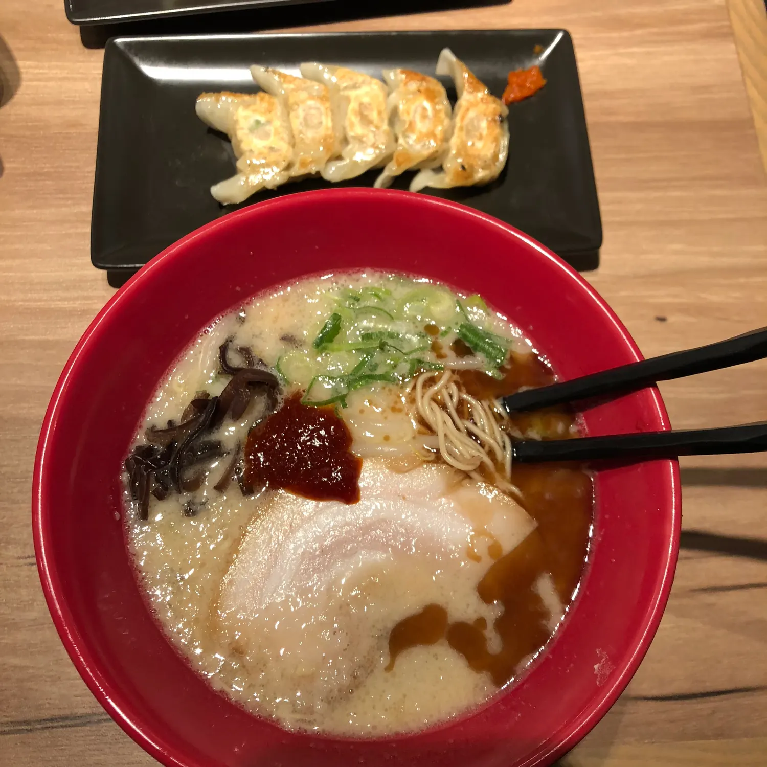 A bowl of noodles in dark broth with a dumpling and greens on top, chopsticks resting on the rim, and a small plate of fried dumplings behind it.