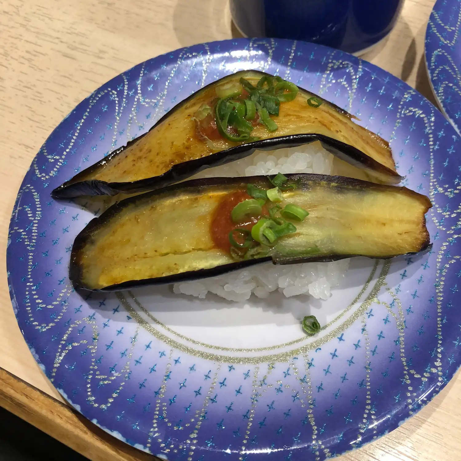 Two halves of grilled aubergine with spring onion and a dab of sauce over rice on a blue patterned plate.