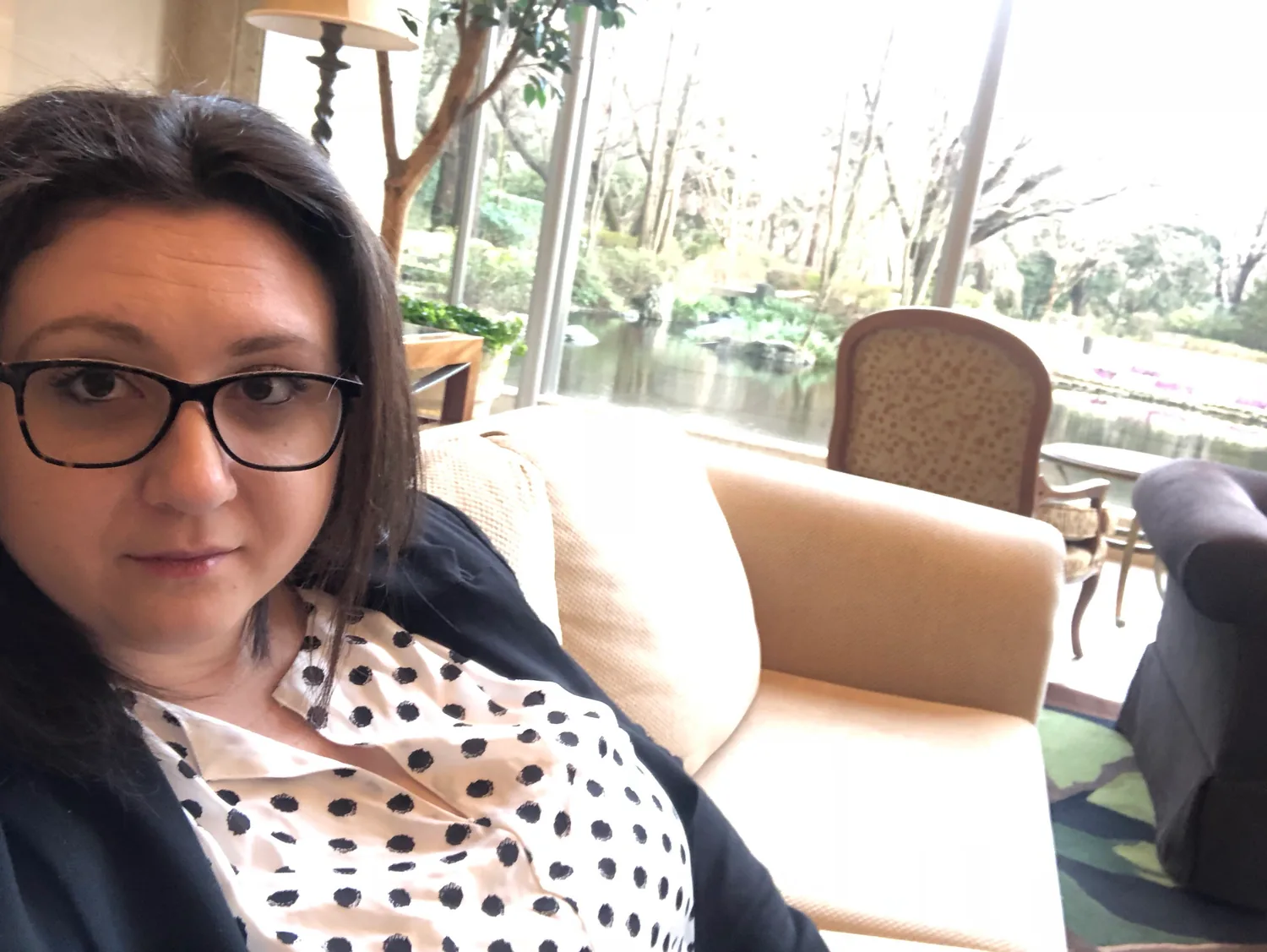 A woman in glasses and a spotted blouse on a sofa beside a tall window, trees and water beyond the glass.