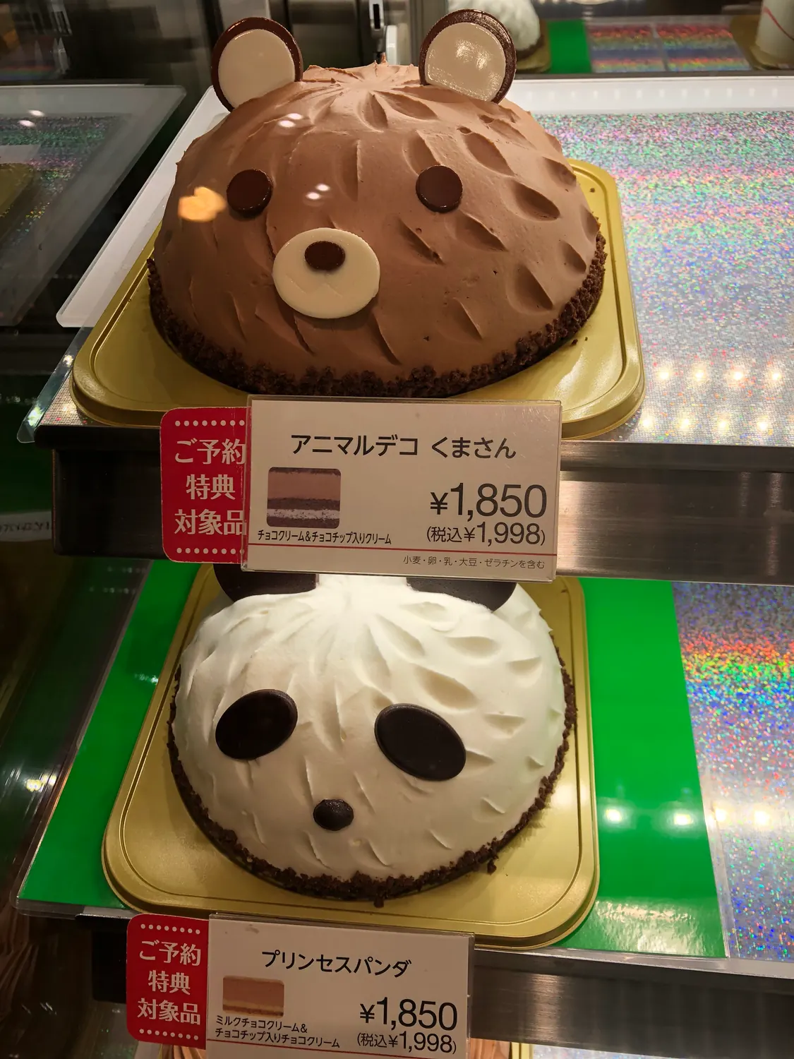 Two animal shaped cakes in paper cases, one chocolate and one cream, with their price cards in front.
