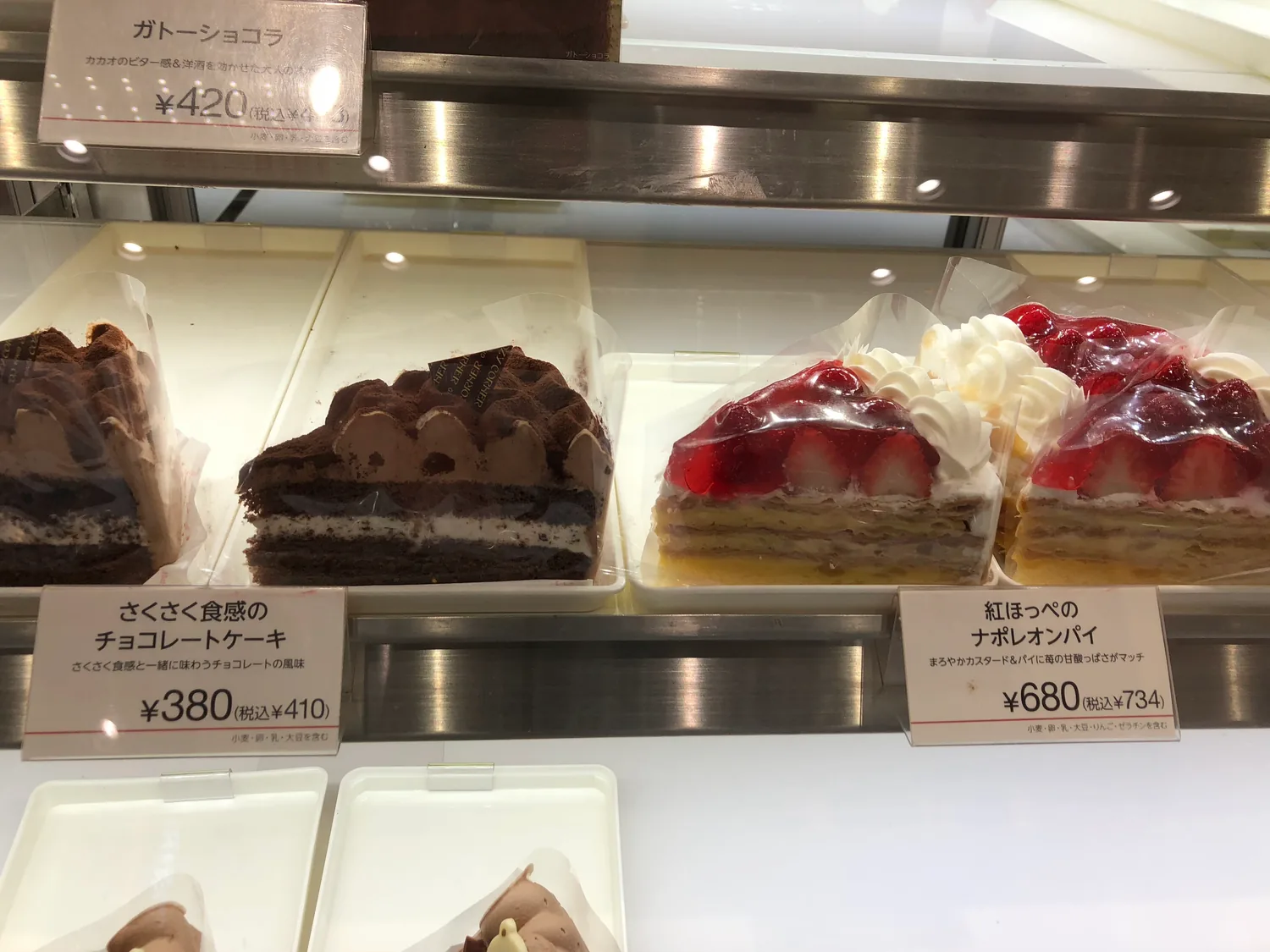 A shop case of chocolate slices and strawberry cream pies with printed labels, price cards along the shelf in front.