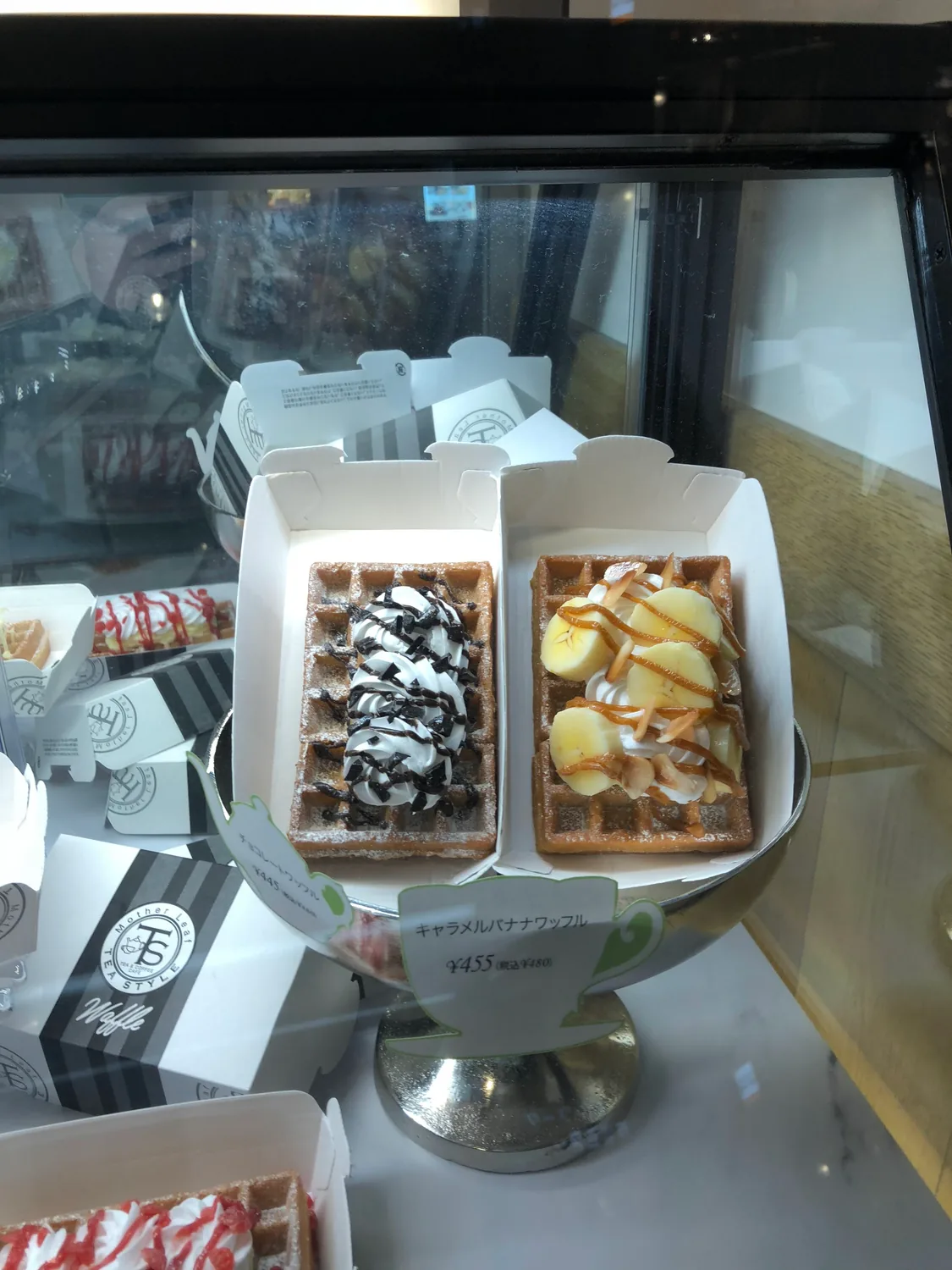 Two waffles in paper boxes on a stand, one under cream and chocolate sauce and one under banana and caramel, a price card in front.