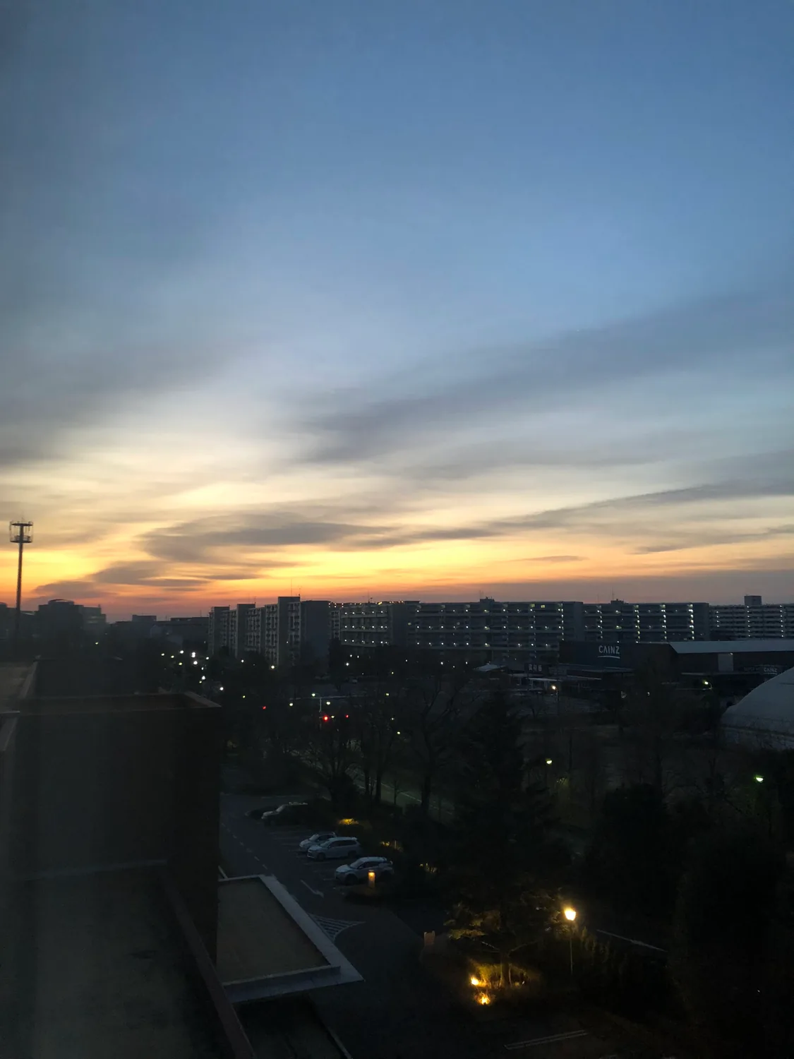 A dawn sky banded with cloud above long lit buildings, bare trees and a car park, seen from a window high above.