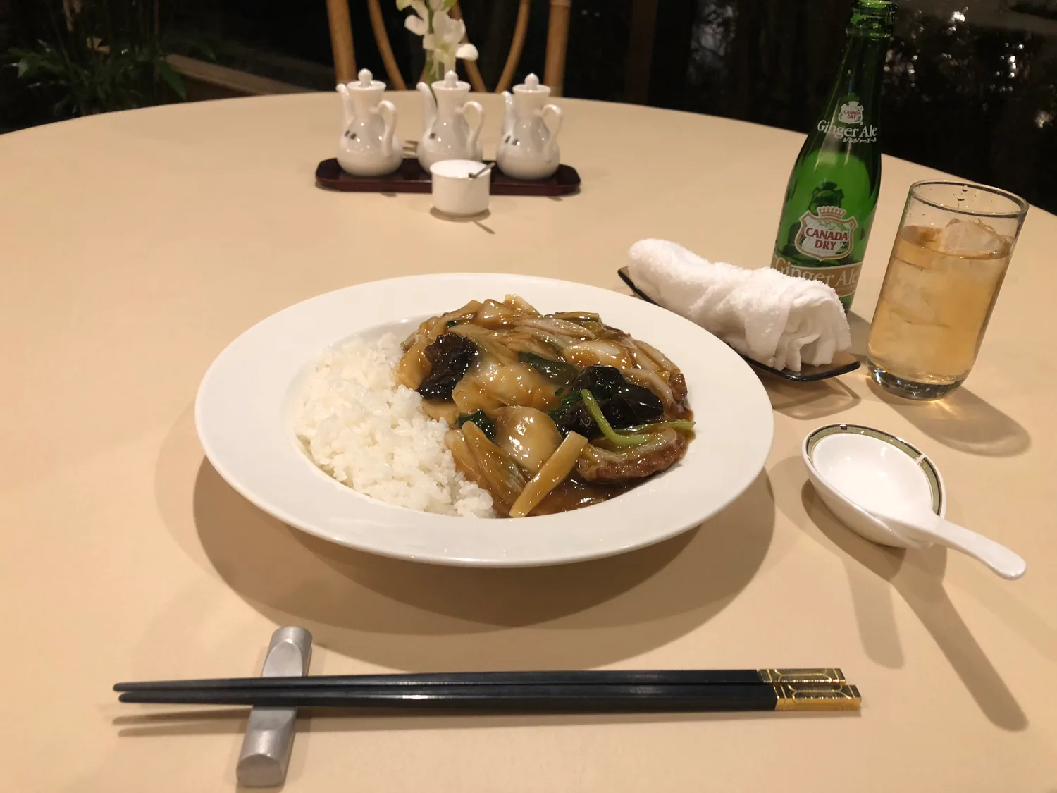 A plate of rice and braised pork with vegetables in sauce, chopsticks beside it, a glass of iced drink and a green bottle at the back.