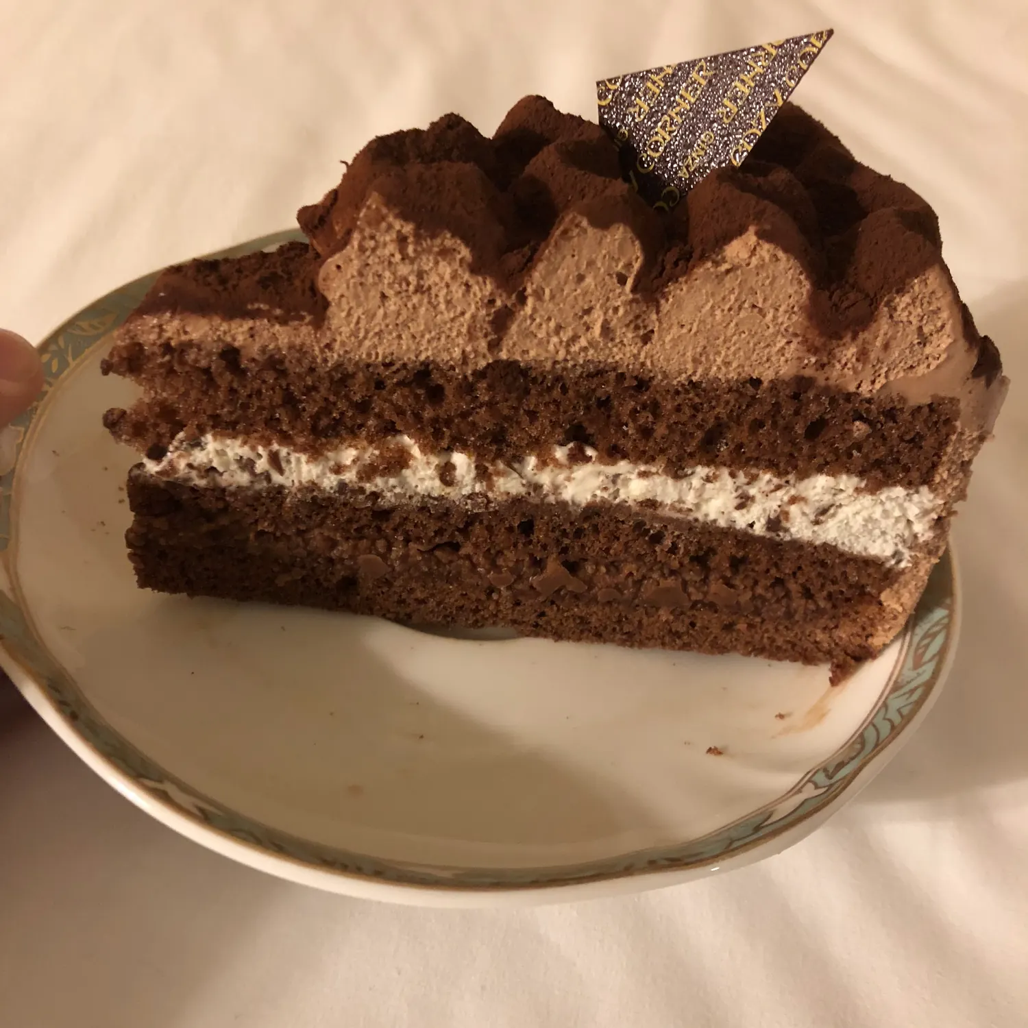 A thick slice of chocolate cake with a layer of cream on a plate, a small printed flag on top.