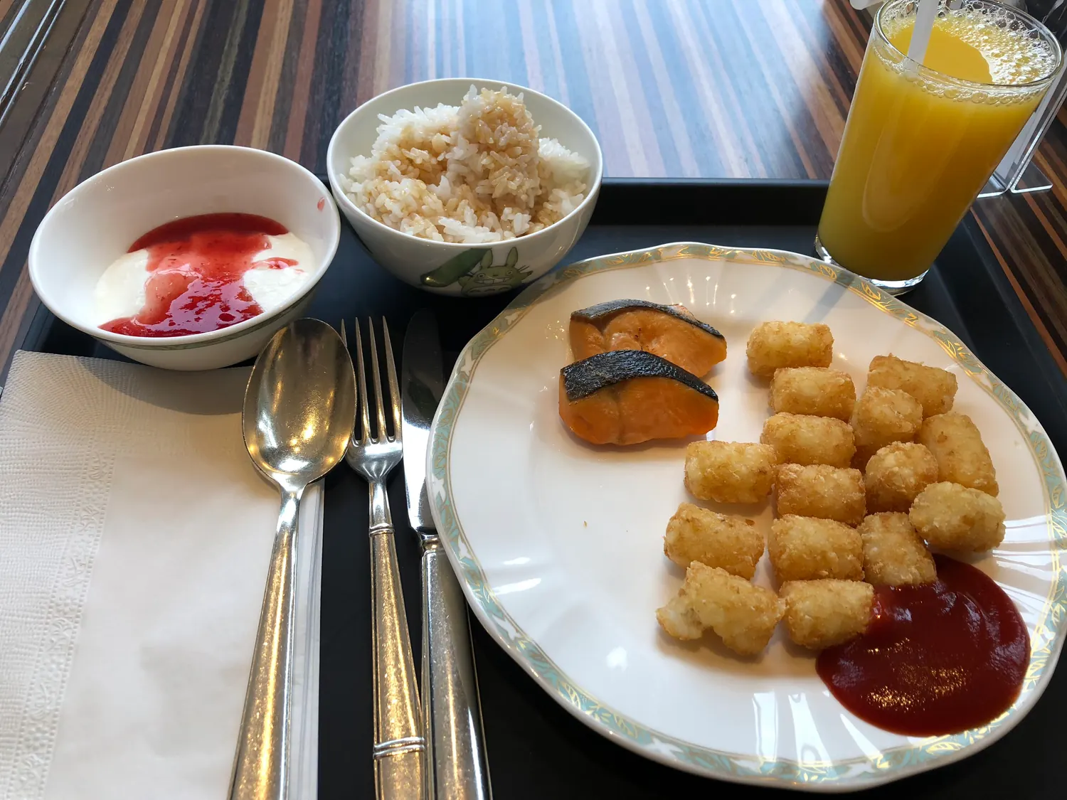A breakfast tray with a bowl of rice, a plate of croquettes and squash with ketchup, a bowl of yoghurt with jam and a glass of juice.