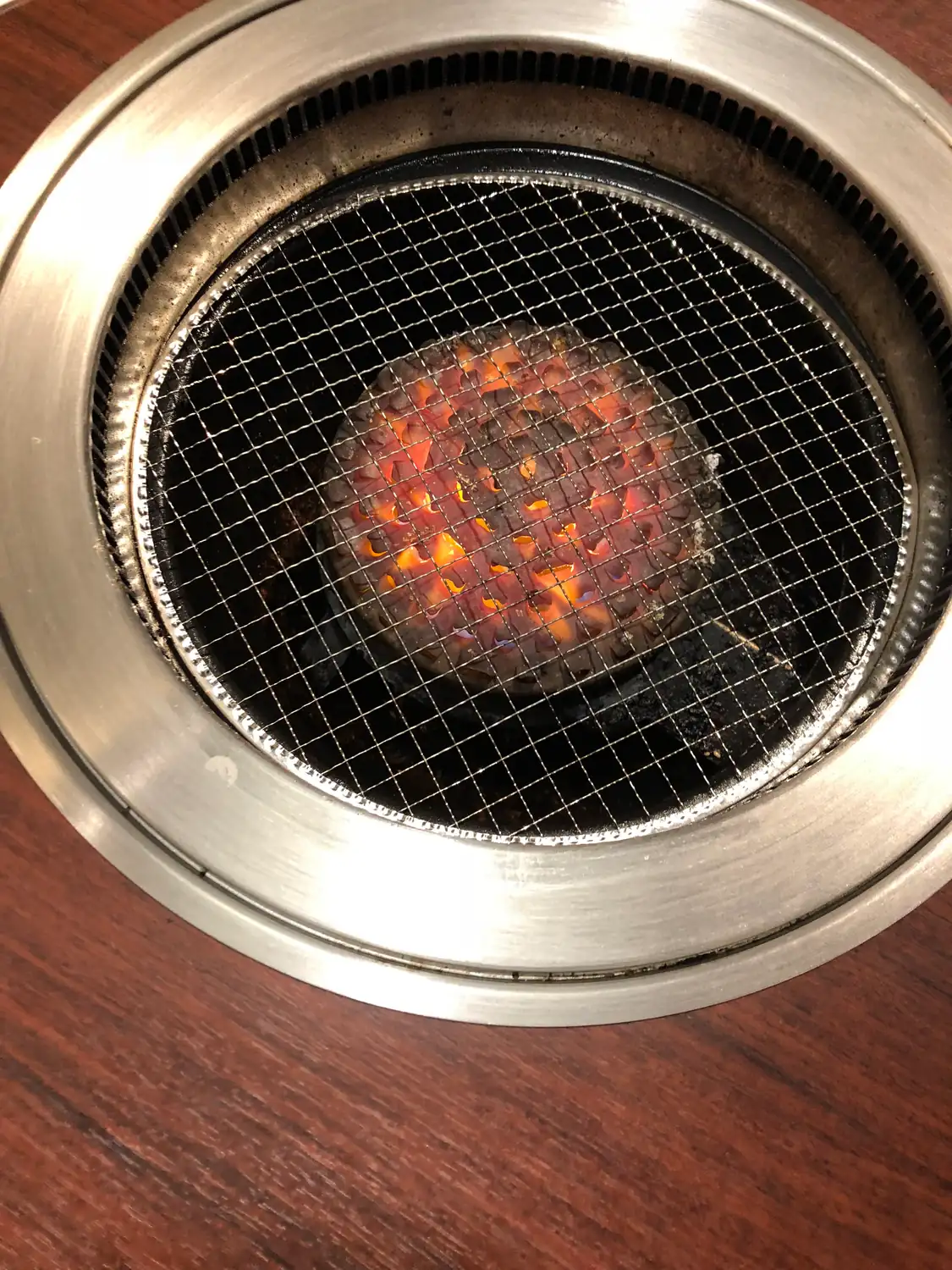 A round grill set into a wooden table, its metal grate over glowing charcoal.