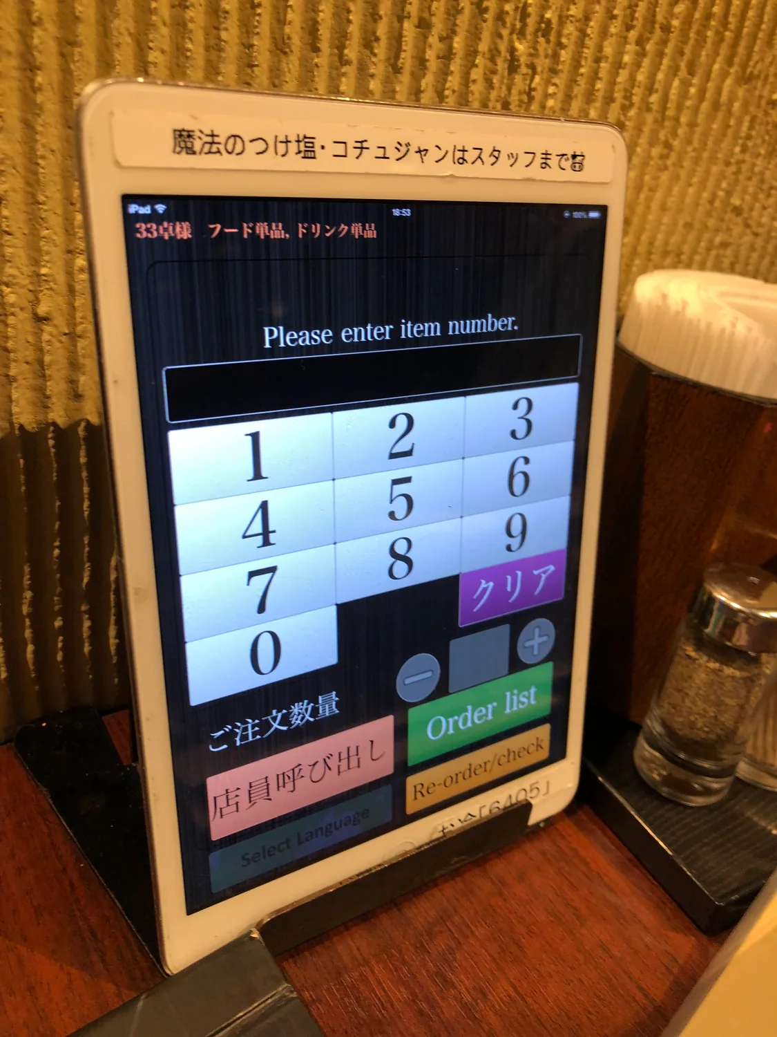 A tablet in a stand on a table showing an ordering screen and a number pad, a wooden stick holder and a shaker beside it.