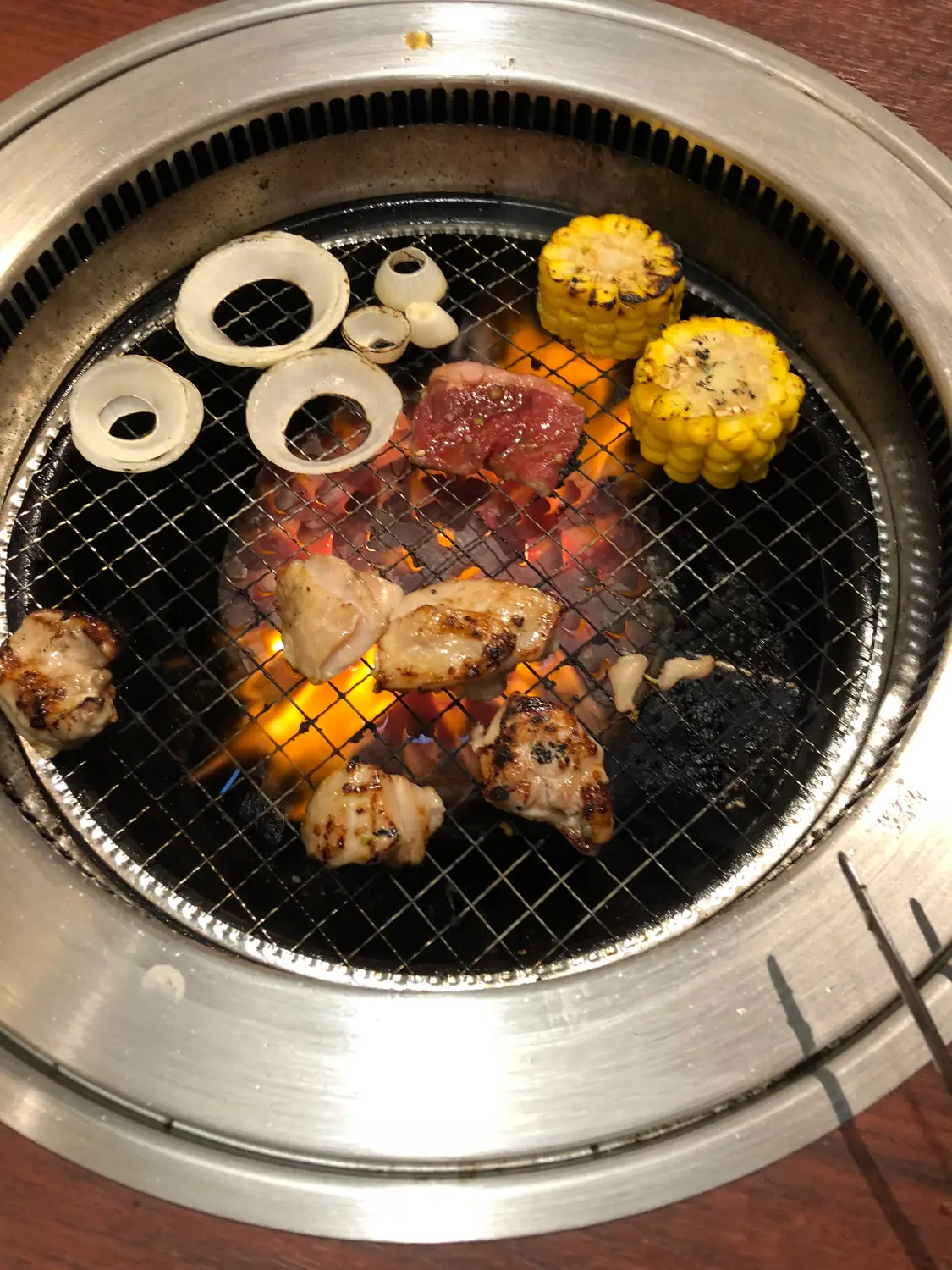 Onion rings, corn cobs and pieces of meat on a table grill over hot coals, the edge of a pair of chopsticks in the corner.