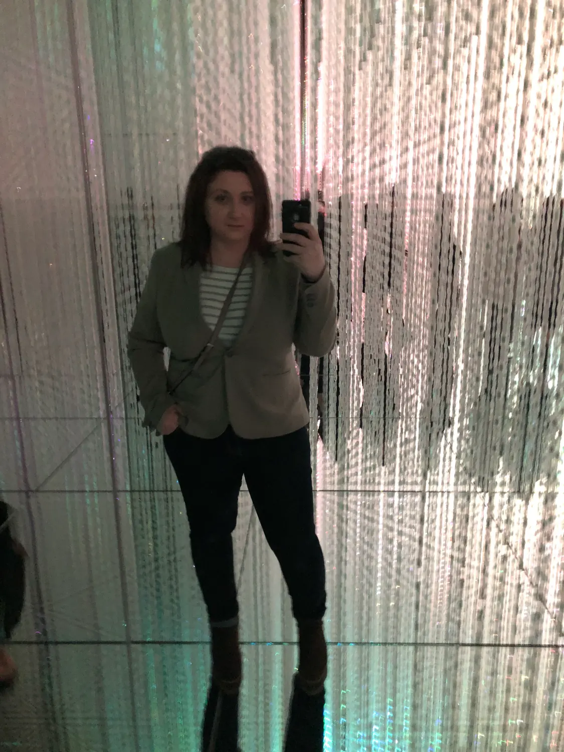 A woman in a green jacket standing in a room lined with mirrored light panels, holding a phone up to take the picture.