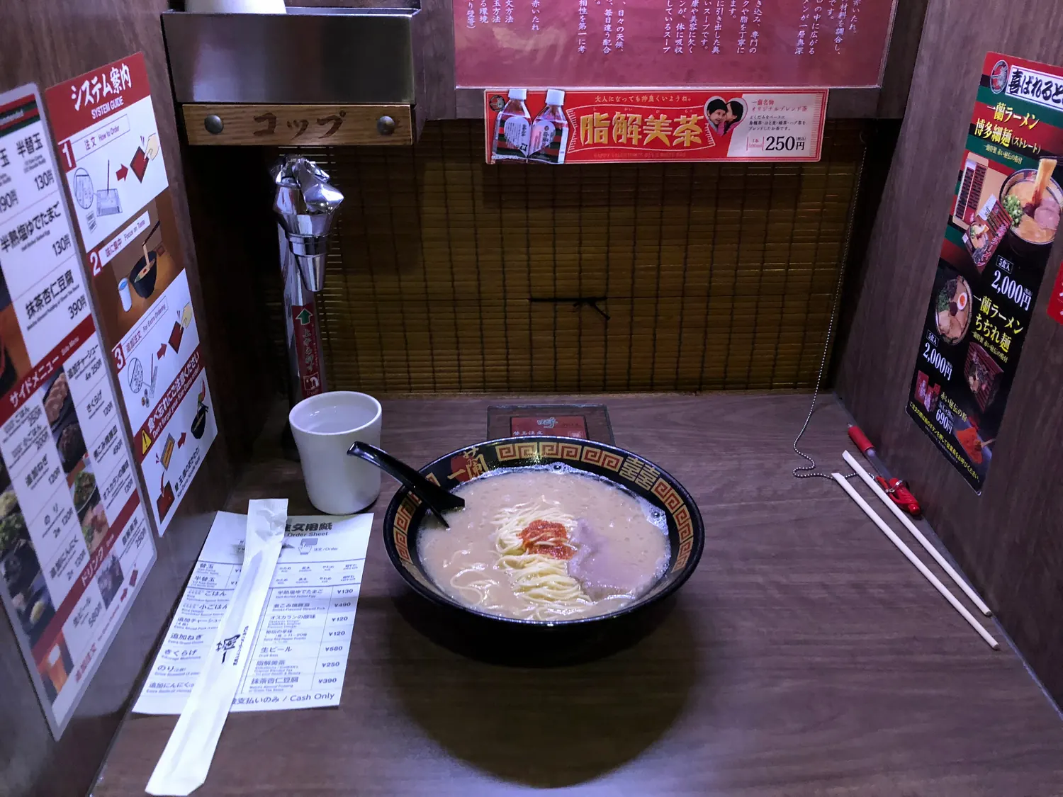 A bowl of noodles on a counter table with chopsticks and a cup beside it, printed notices on the wall behind.