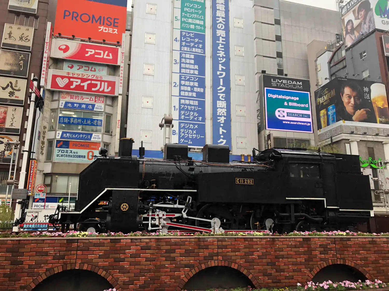 A preserved black steam locomotive standing on a brick arched bridge, with city buildings rising behind it.