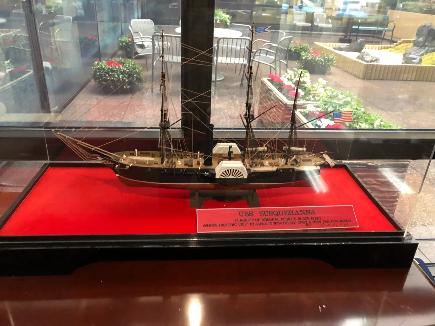 A model sailing ship with three masts in a glass case on a red base, a small printed card in front and a courtyard of seats and plants beyond the glass.
