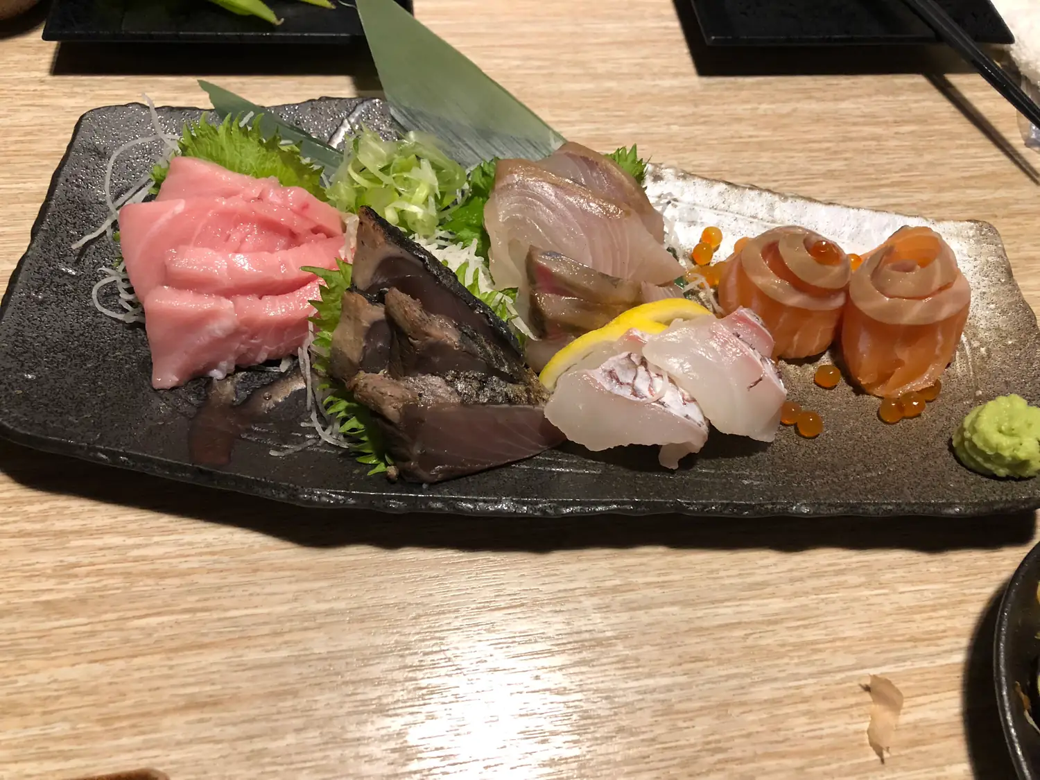 Slices of raw fish and seafood rolls on a dark slate plate with shredded garnish and a piece of wasabi.