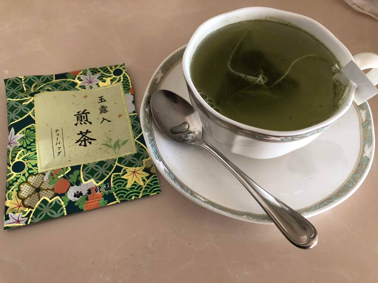 A cup of green tea on a saucer with its spoon, beside a patterned packet of tea leaves on a tray.