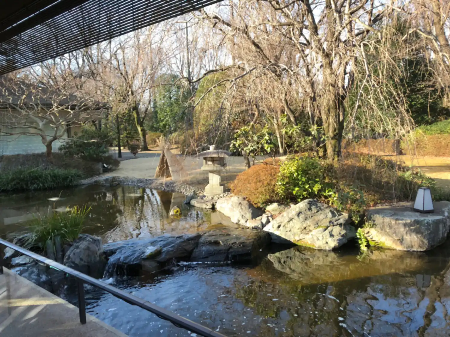 A garden pond with a small fall over its stones, a stone lantern and bare trees around it, seen from under a slatted roof.